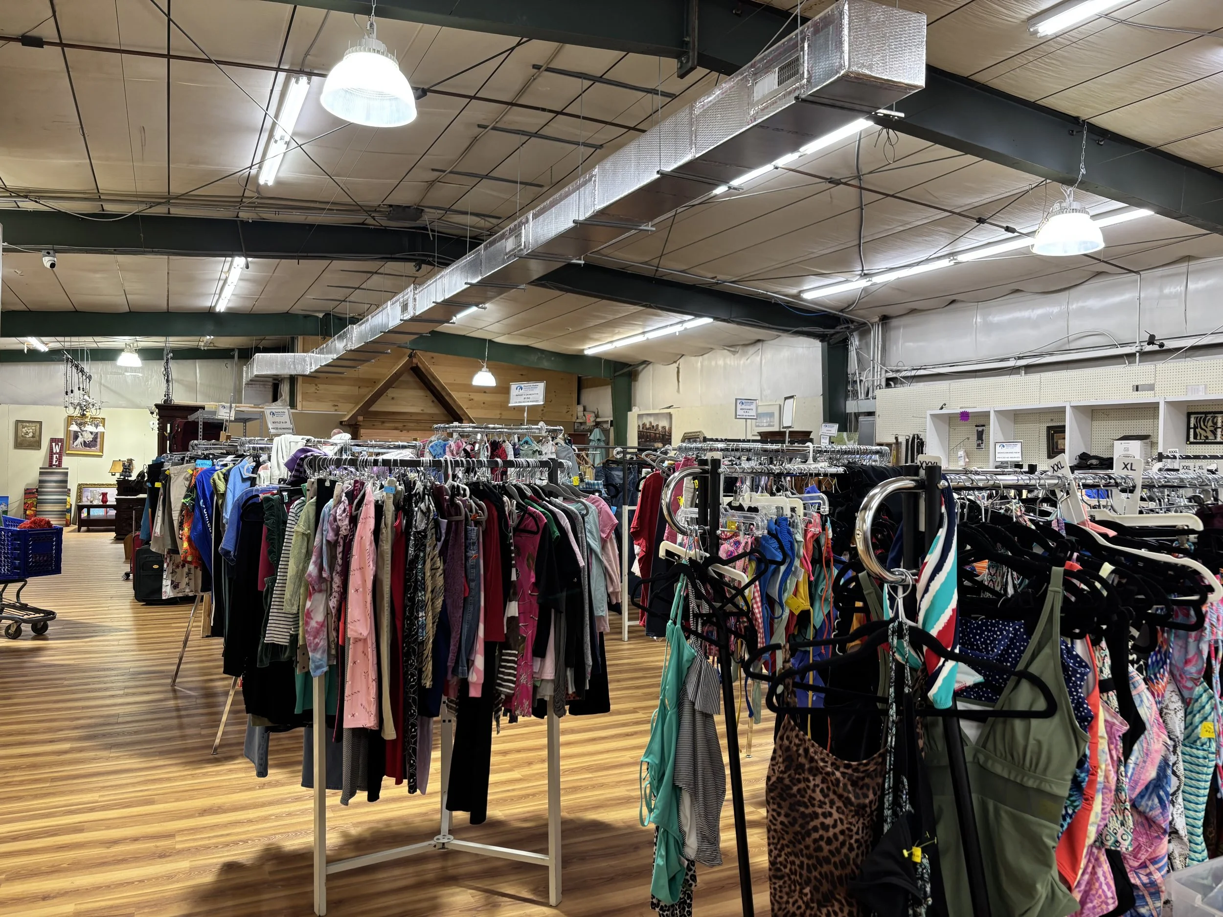 Clothing racks filled with various clothes inside a store, with a wooden floor, ceiling lights, and a wall with framed pictures.