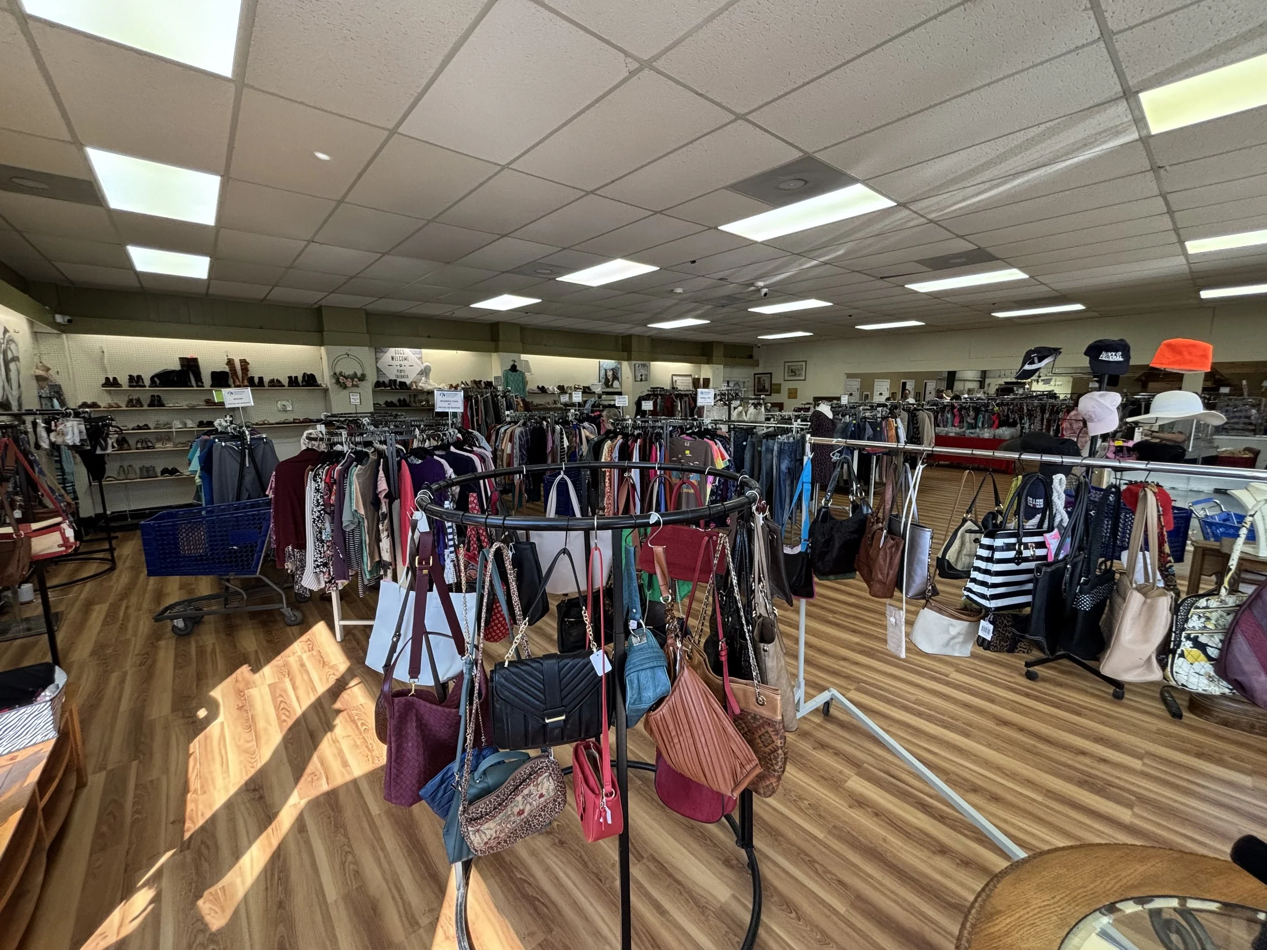 Interior of a thrift store with shelves of shoes, racks of clothing, and display tables filled with handbags and accessories.