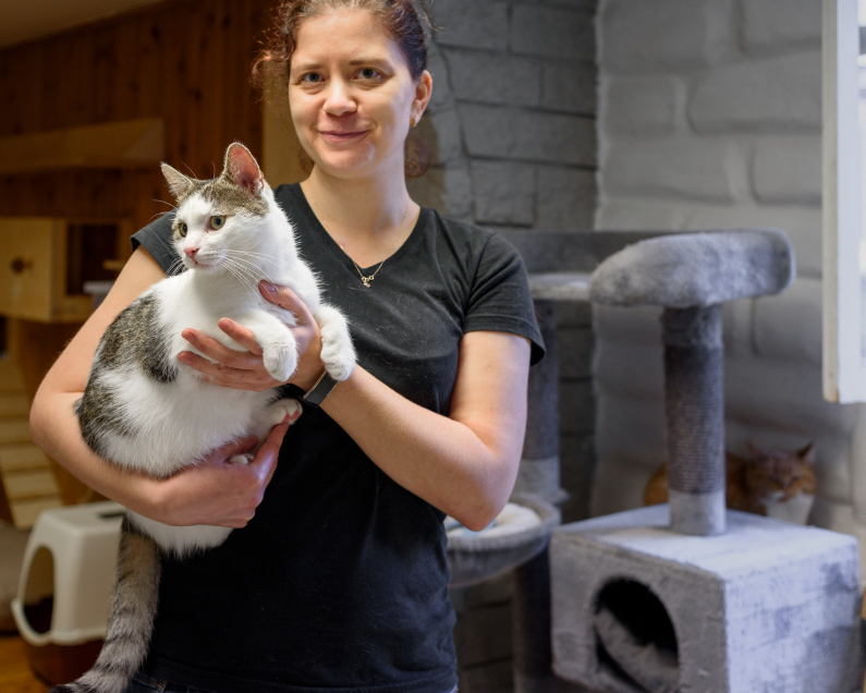A woman holding a white and gray tabby cat indoors with a cat tree in the background.
