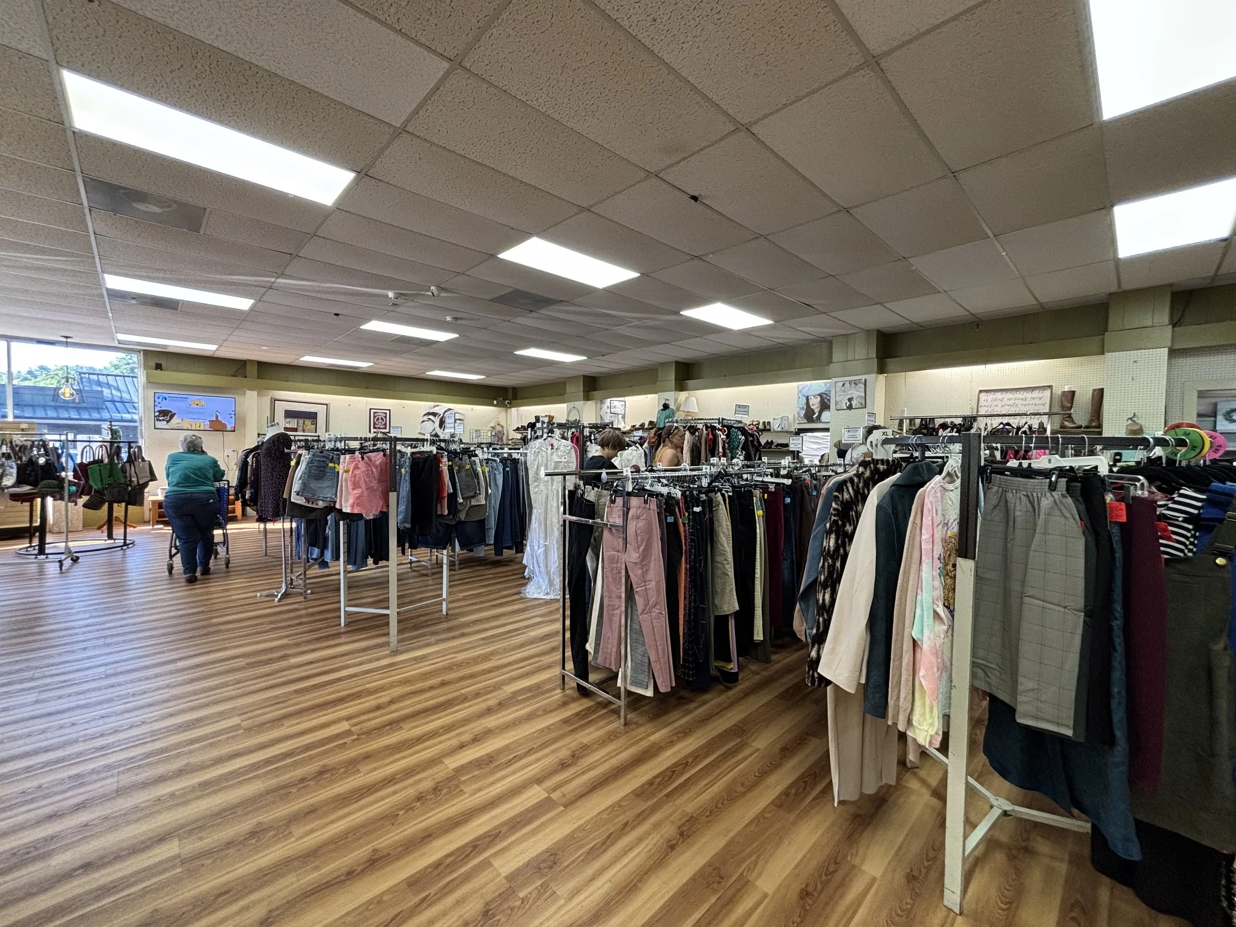 Inside a thrift store or clothing resale shop with multiple racks of clothing, some shelves, and a few shoppers browsing. The ceiling has fluorescent lights, and the floor is wood-like.