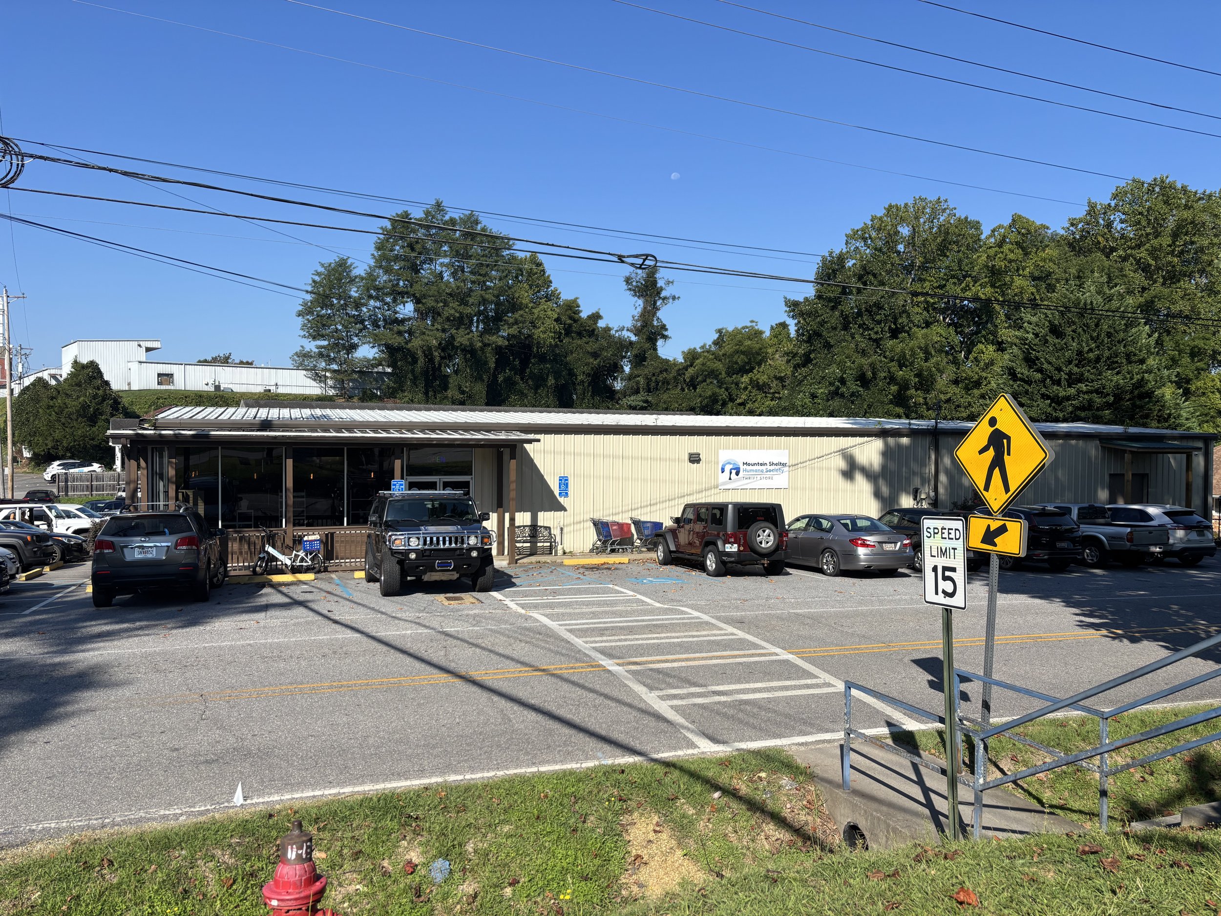 A parking lot with cars, a building with a sign for Mountain Shelter Humane Society Thrift Store, road signs including a pedestrian crossing and speed limit, and trees in the background.