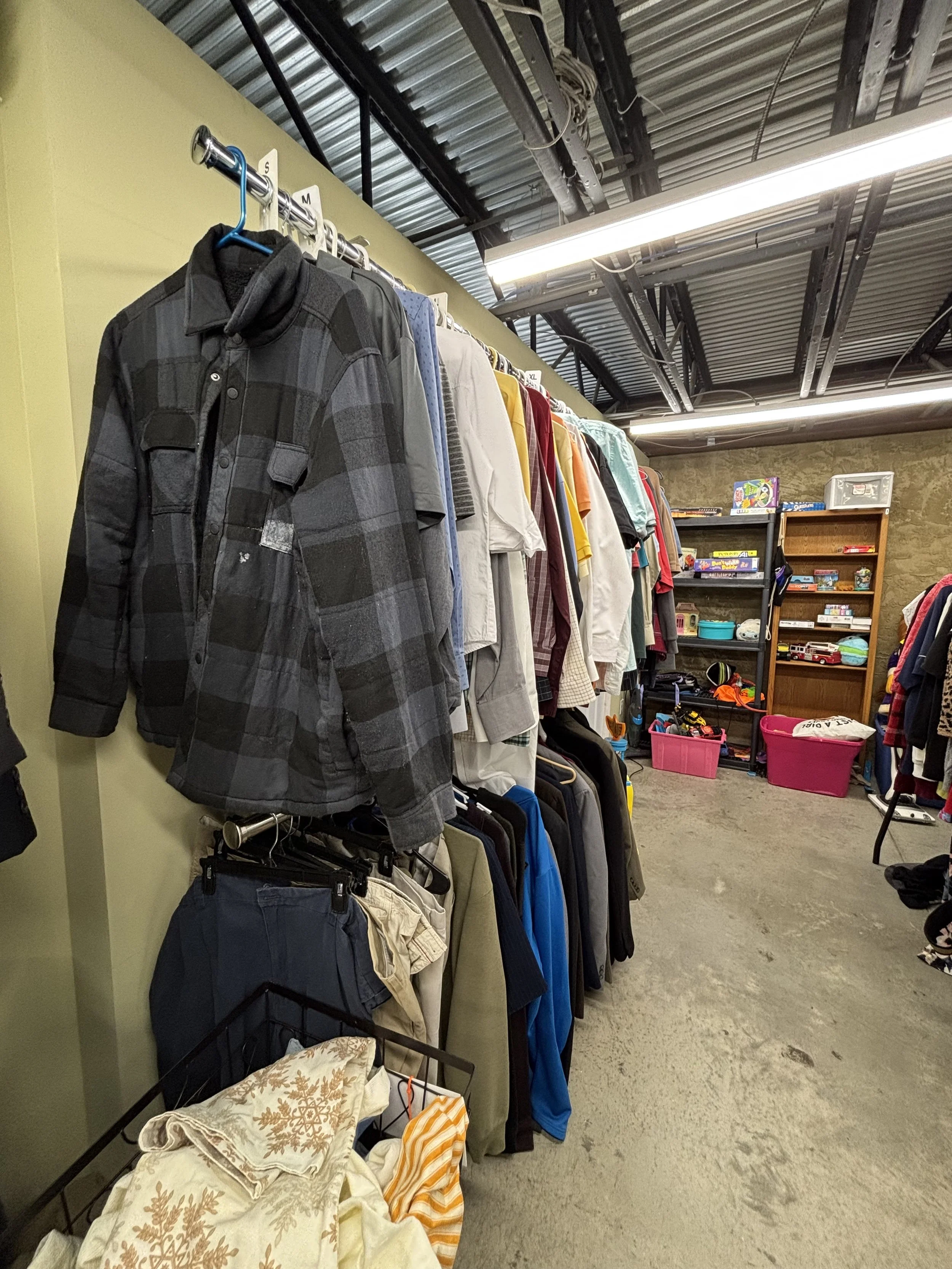 Clothing rack with various shirts and jackets in a thrift store or secondhand shop. Shelves with toys, books, and storage bins in the background.
