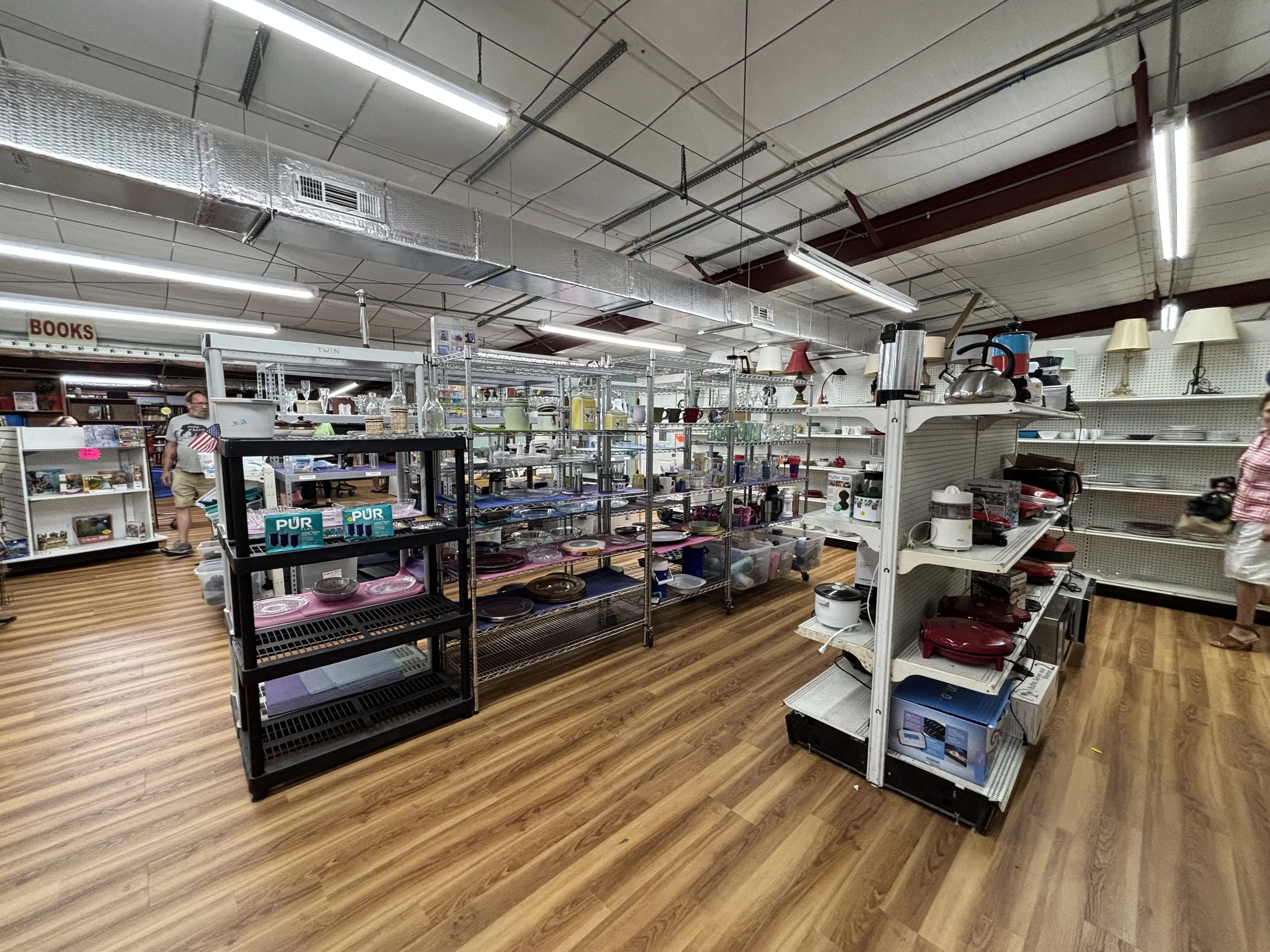 Interior of a thrift store or flea market with shelves filled with various household items such as lamps, dishes, and small appliances. Fluorescent lights and exposed ceiling ductwork are visible.