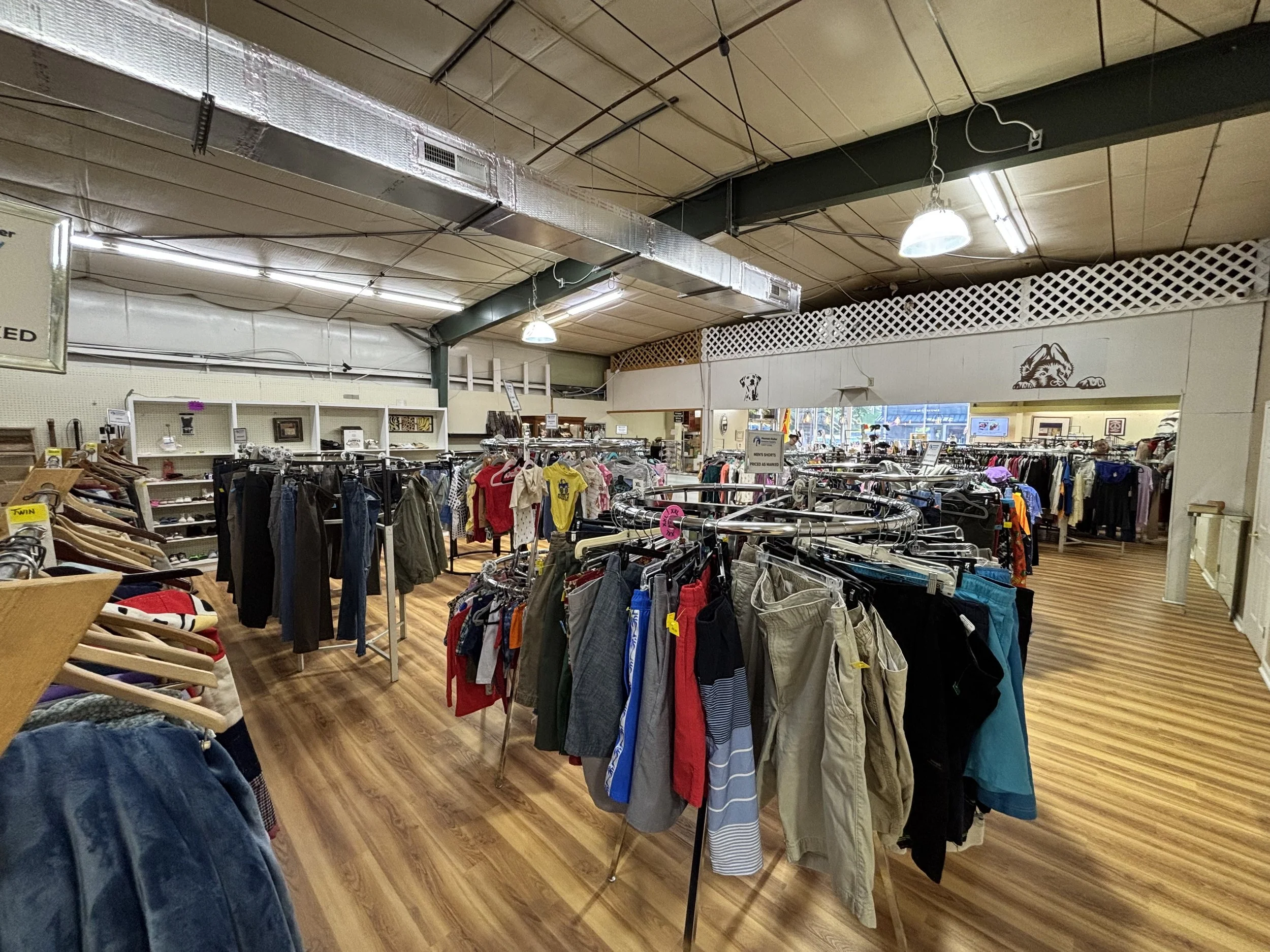 Clothing racks filled with various casual and athletic wear inside a thrift or secondhand store, with a wooden floor and overhead lighting.