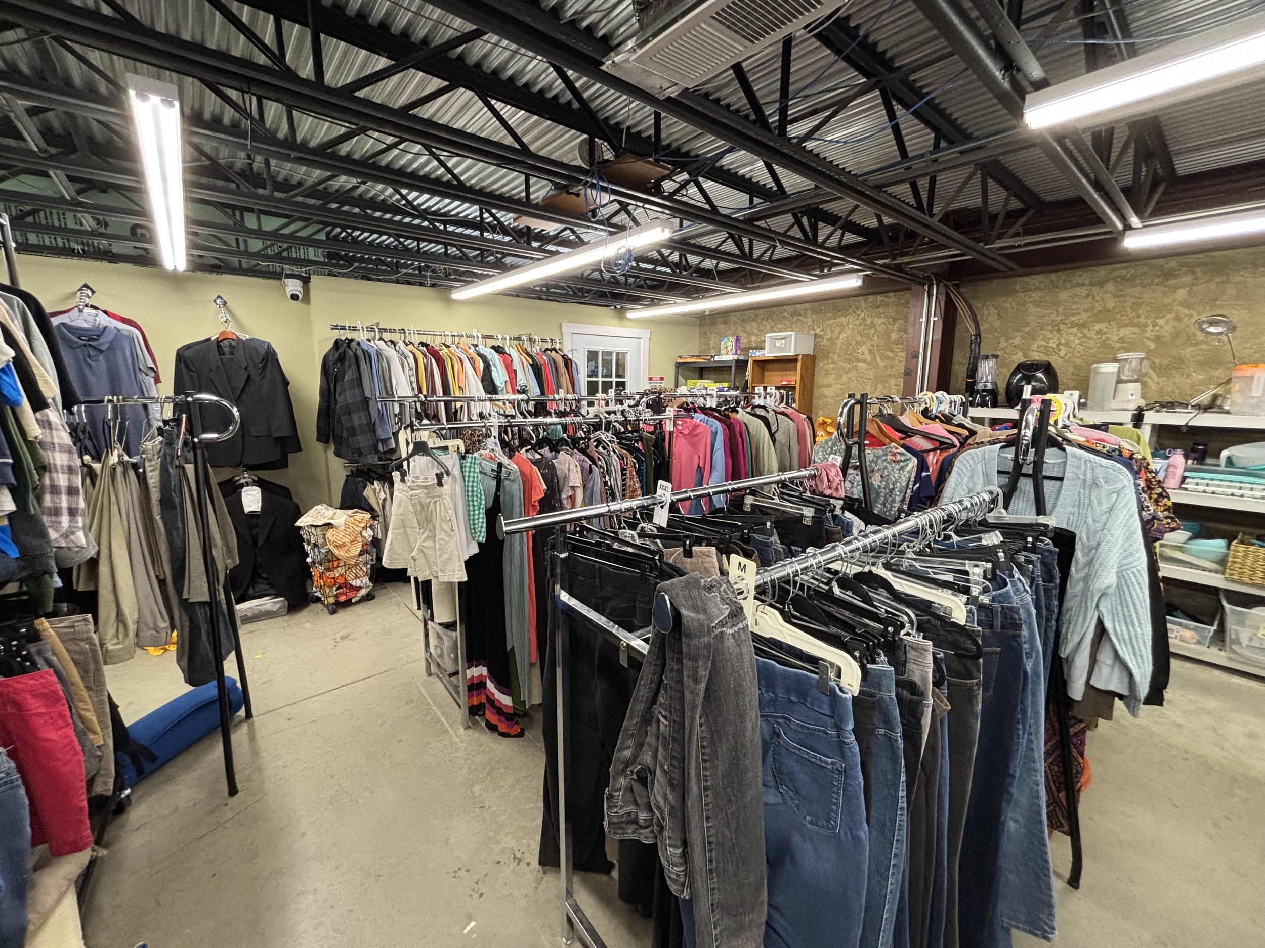 Inside a thrift store or secondhand clothing shop with racks of clothes and apparel for sale, fluorescent lighting, and a concrete floor.
