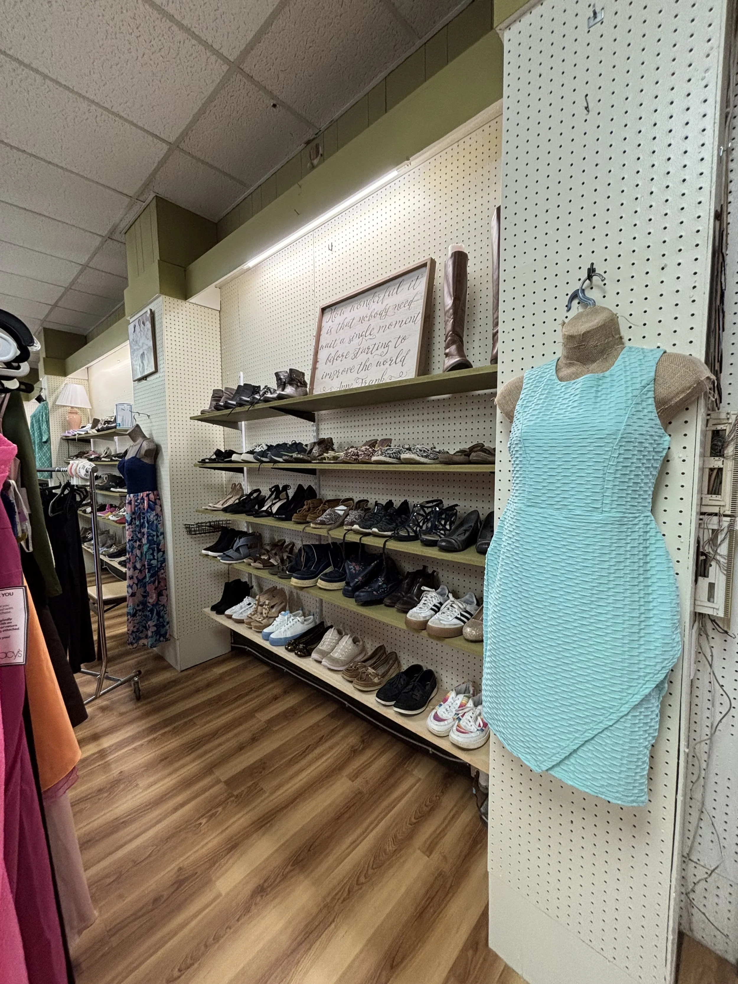 Store display with shoes on shelves, a mannequin wearing a sleeveless turquoise textured dress, and a framed quote about kindness and improvement.