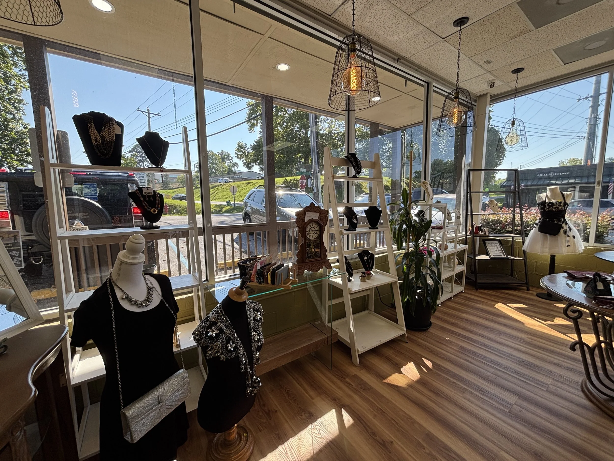 Inside a jewelry or clothing store with large glass windows, mannequins displaying necklaces and accessories, and pendant lights hanging from the ceiling. Outside view of parked cars and trees.