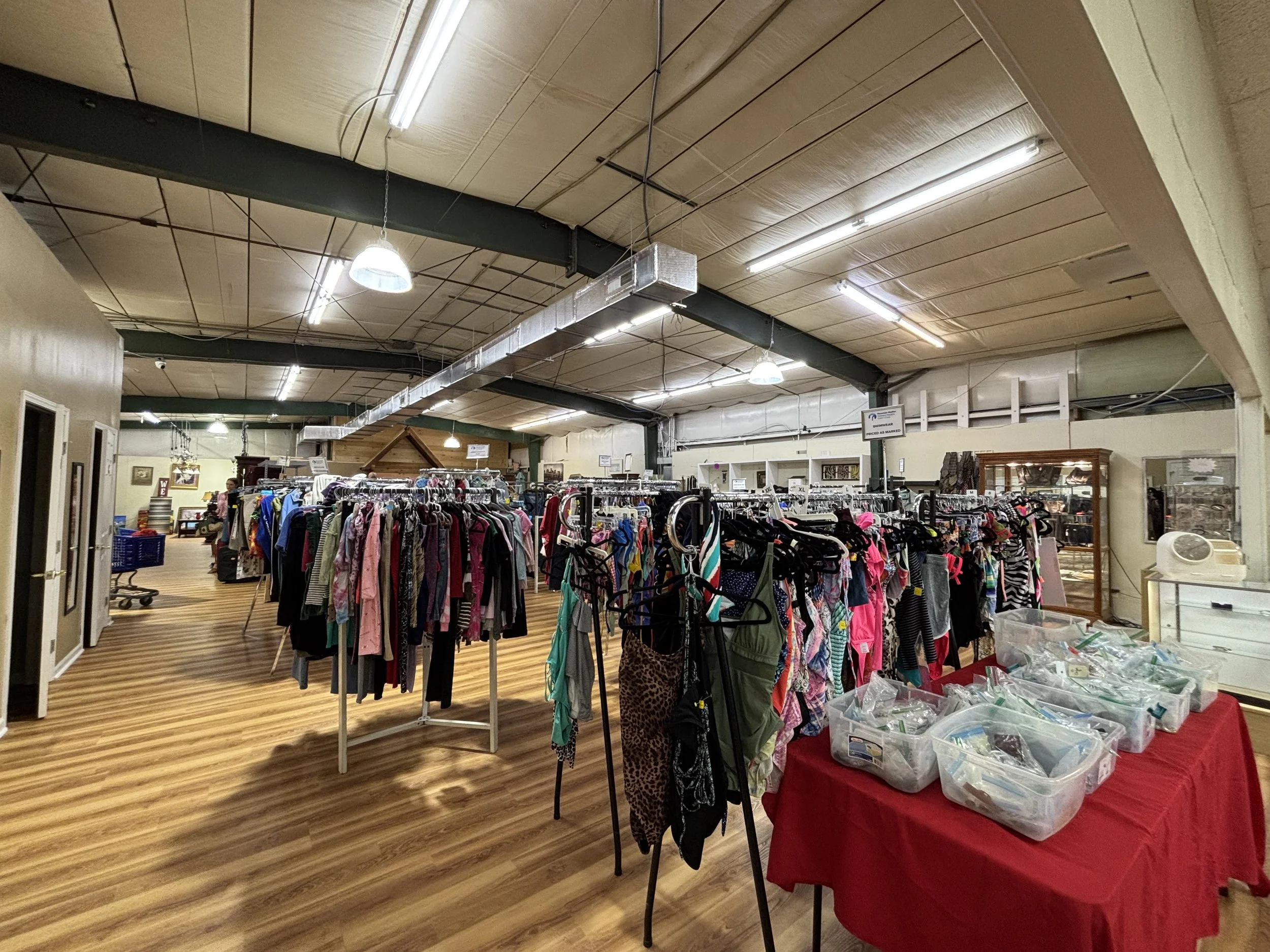 The image depicts the interior of a thrift store or second-hand shop, with clothing racks filled with various garments, a table with clear plastic containers holding small items, and signs hanging from the ceiling. The ceiling has exposed beams, duct