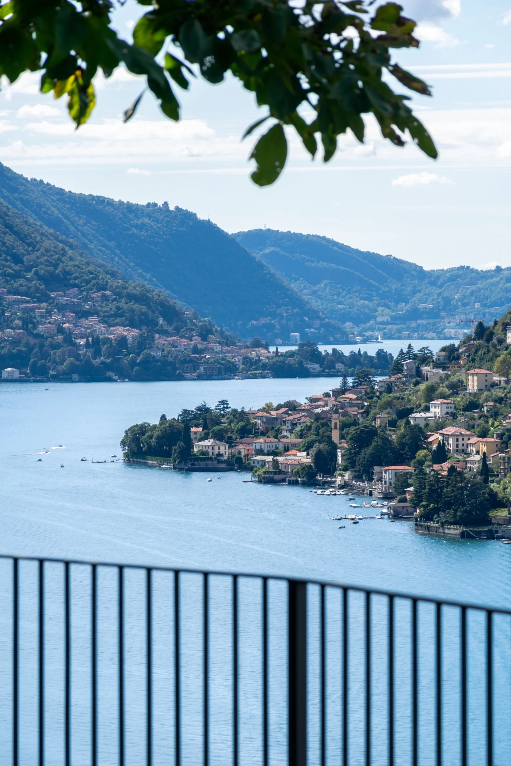 Vista panoramica sul lago di Como e sulle montagne, incorniciata da una recinzione in ferro e rami di albero.