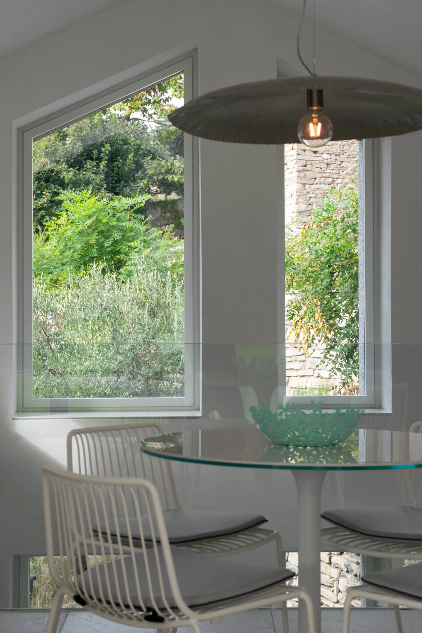 Sala da pranzo moderna con tavolo in vetro, lampada Brass Gervasoni e vista sul giardino attraverso finestre asimmetriche.