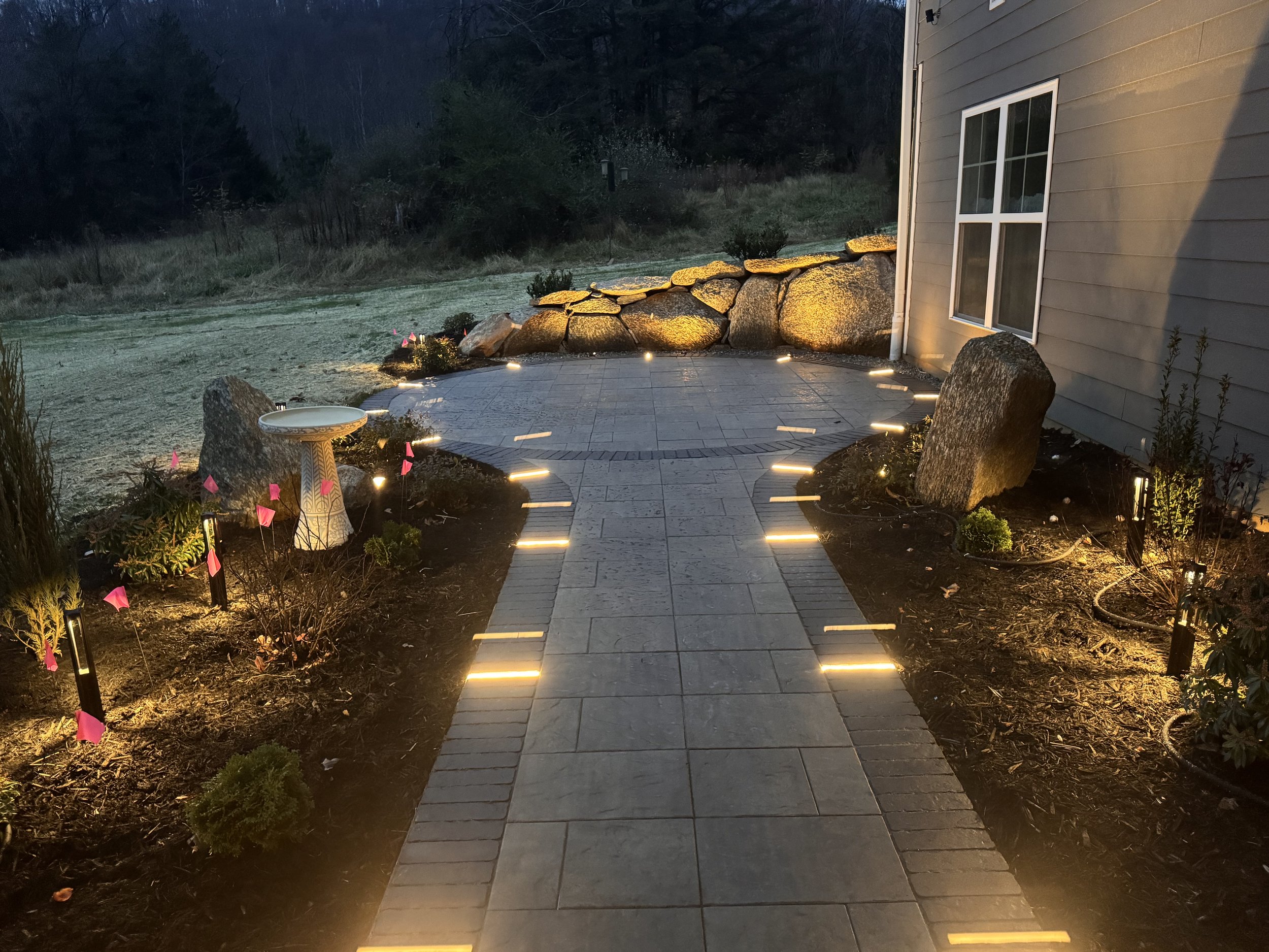 Nighttime backyard scene featuring a paved walkway with embedded lights leading to a stone patio, surrounded by landscaped garden beds with small plants and outdoor lighting, next to a beige house with a window, and large rocks in the background.