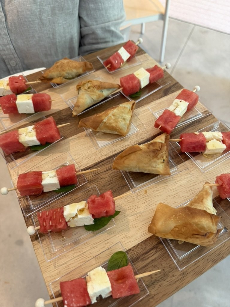 Skewers with watermelon and cheese alternating over a green leaf, and samosas, all arranged on a wooden tray.