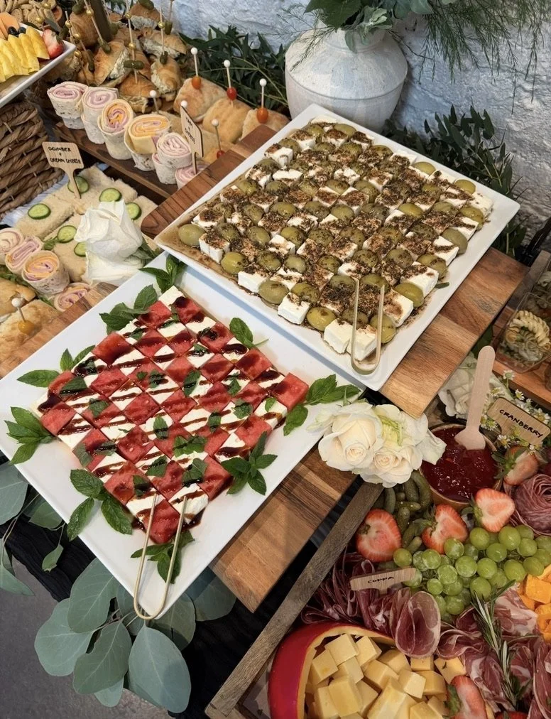 A buffet table with assorted desserts and fresh fruit. The top rack has mini sandwiches and rolled pastries, while the large white trays in front contain caprese skewers with tomatoes, mozzarella, basil, and balsamic glaze, along with grape and strawberry skewers, and other assorted fruit and cheese platters.