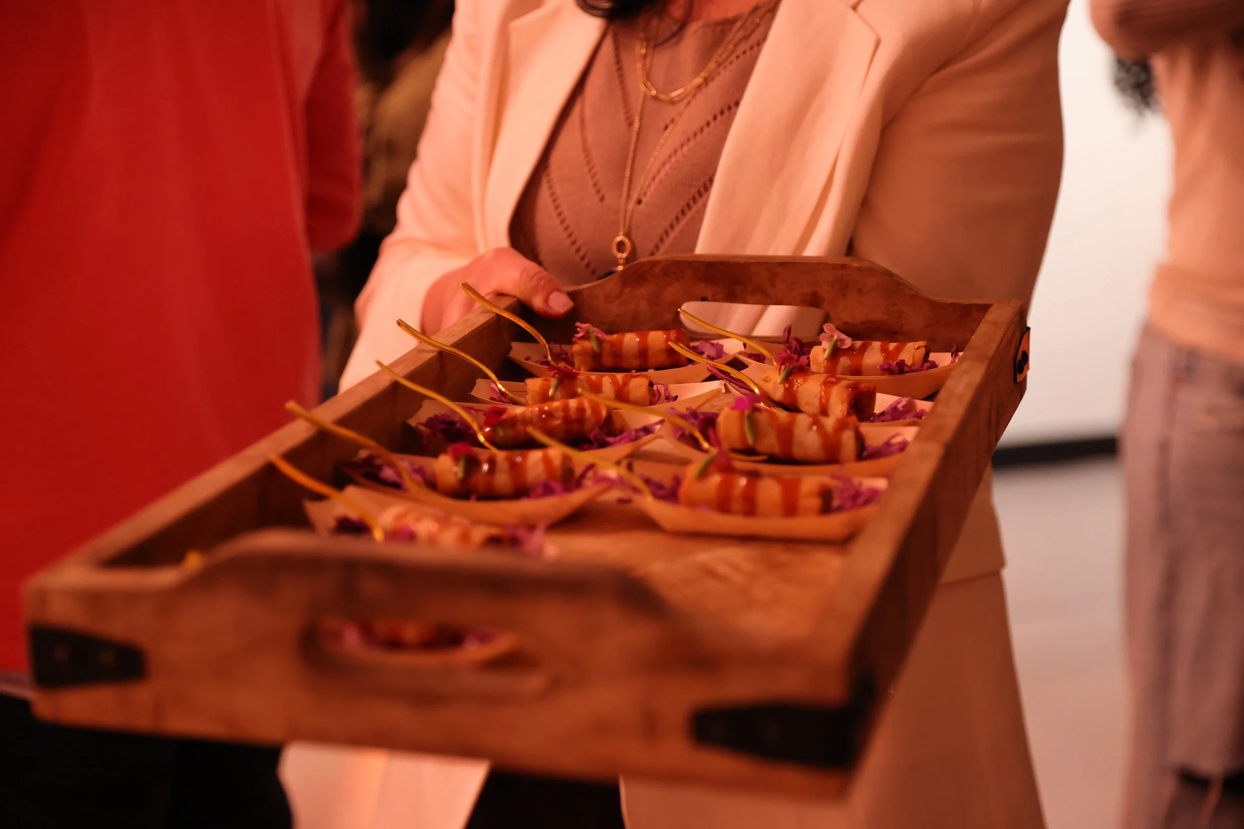 A person wearing a light-colored blazer and gold jewelry holding a wooden tray with appetizers that have orange and white sauces, garnished with purple edible flowers.