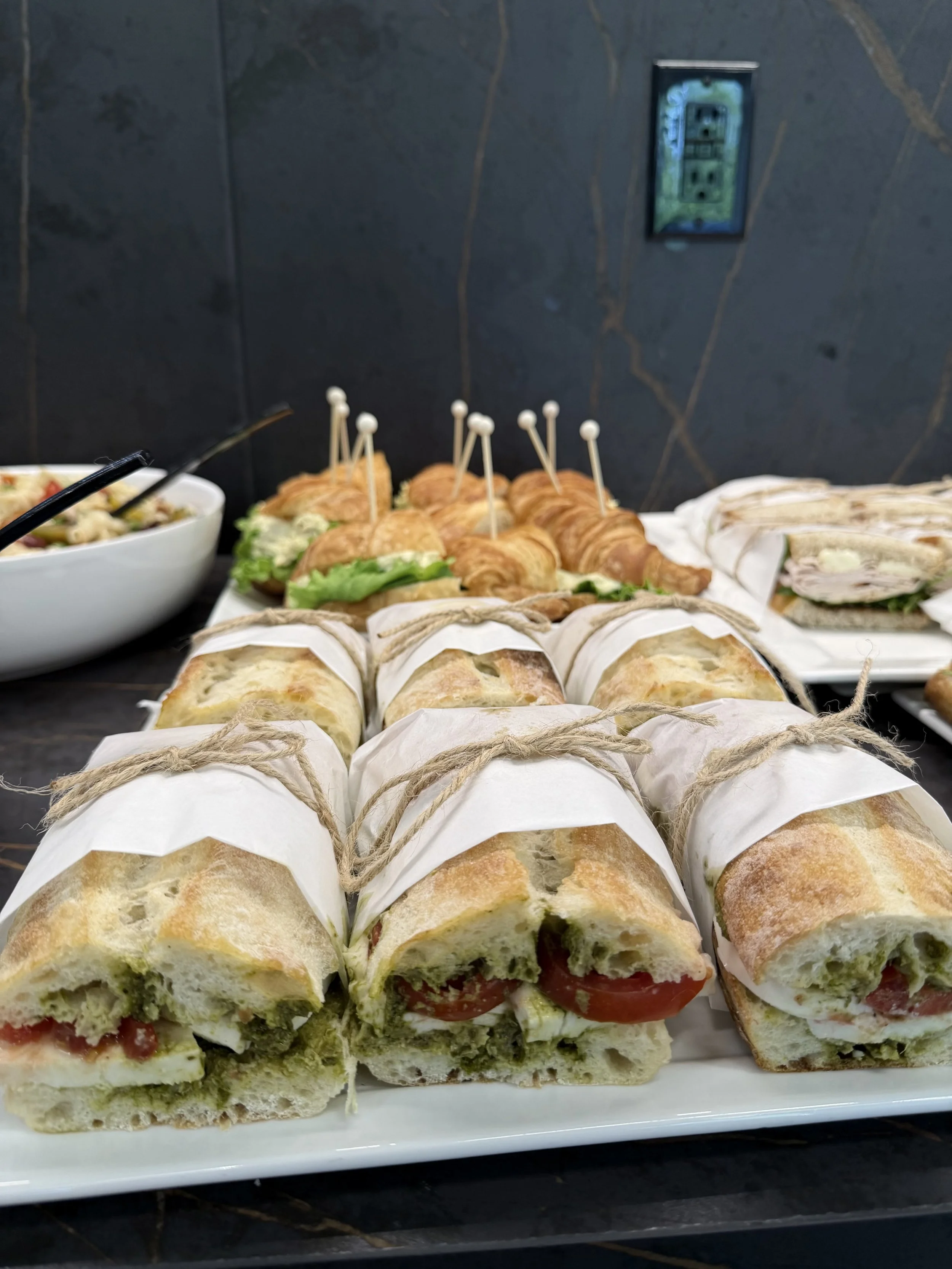 A platter of assorted sandwiches and wraps, including mini croissant sandwiches and tortilla wraps, displayed on a white tray against a dark tiled wall.