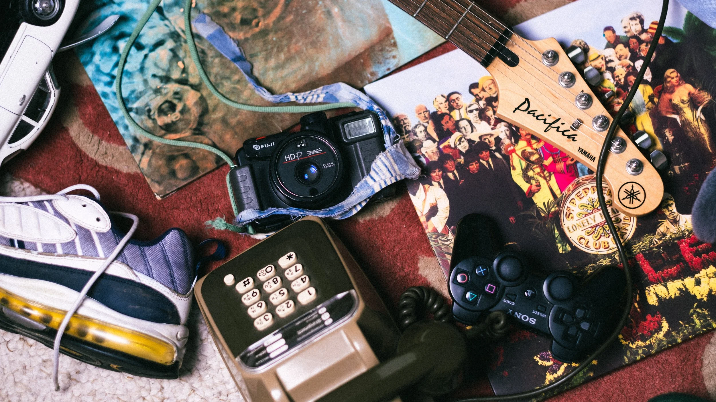 Divers objets sur un tapis rouge, notamment un appareil photo Fuji, une paire de chaussures de sport, une guitare Yamaha, une manette de jeu PlayStation, un téléphone fixe, un casque audio, un disque vinyle ainsi qu'une affiche ou un magazine avec plusieurs personnes en couleur.