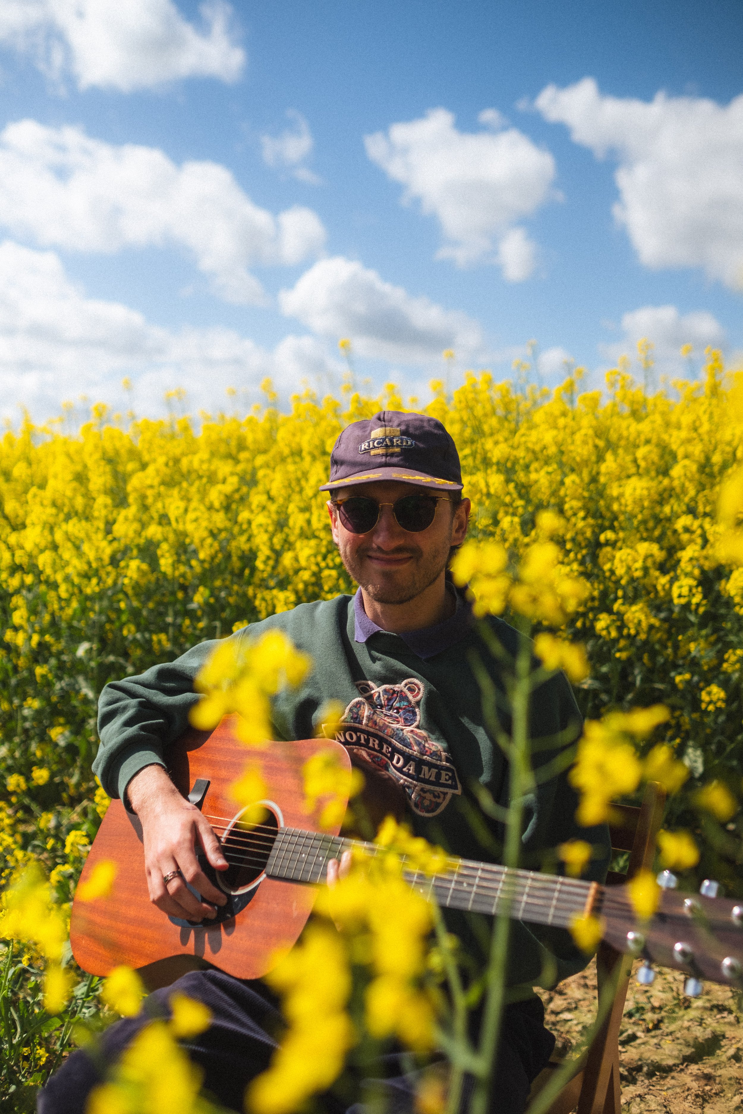 Un homme joue de la guitare dans un champ de fleurs jaunes sous un ciel bleu avec quelques nuages.