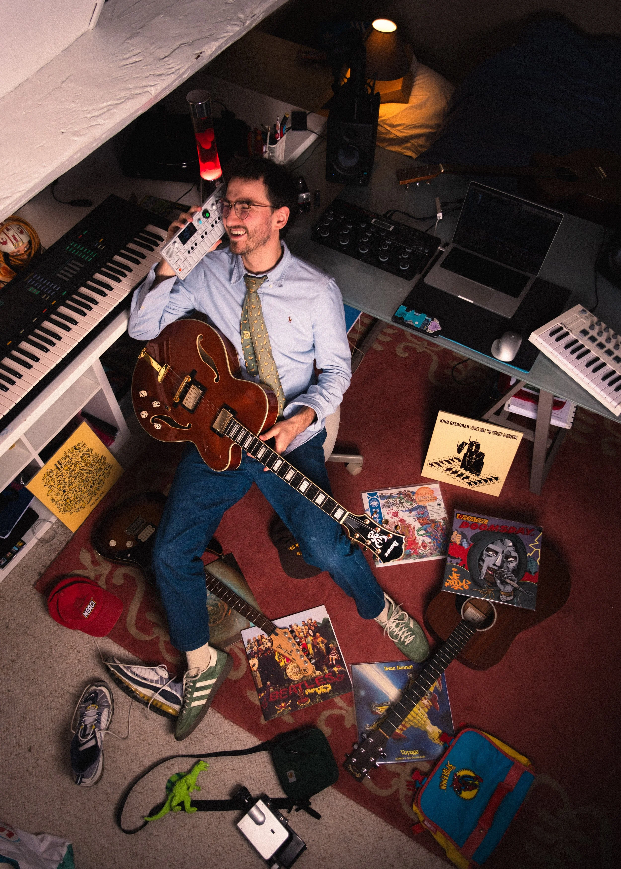 Un homme souriant, jouant de la guitare et parlant au téléphone dans un studio de musique, entouré de vinyles, appareils électroniques et même une enceinte de haute fidélité.