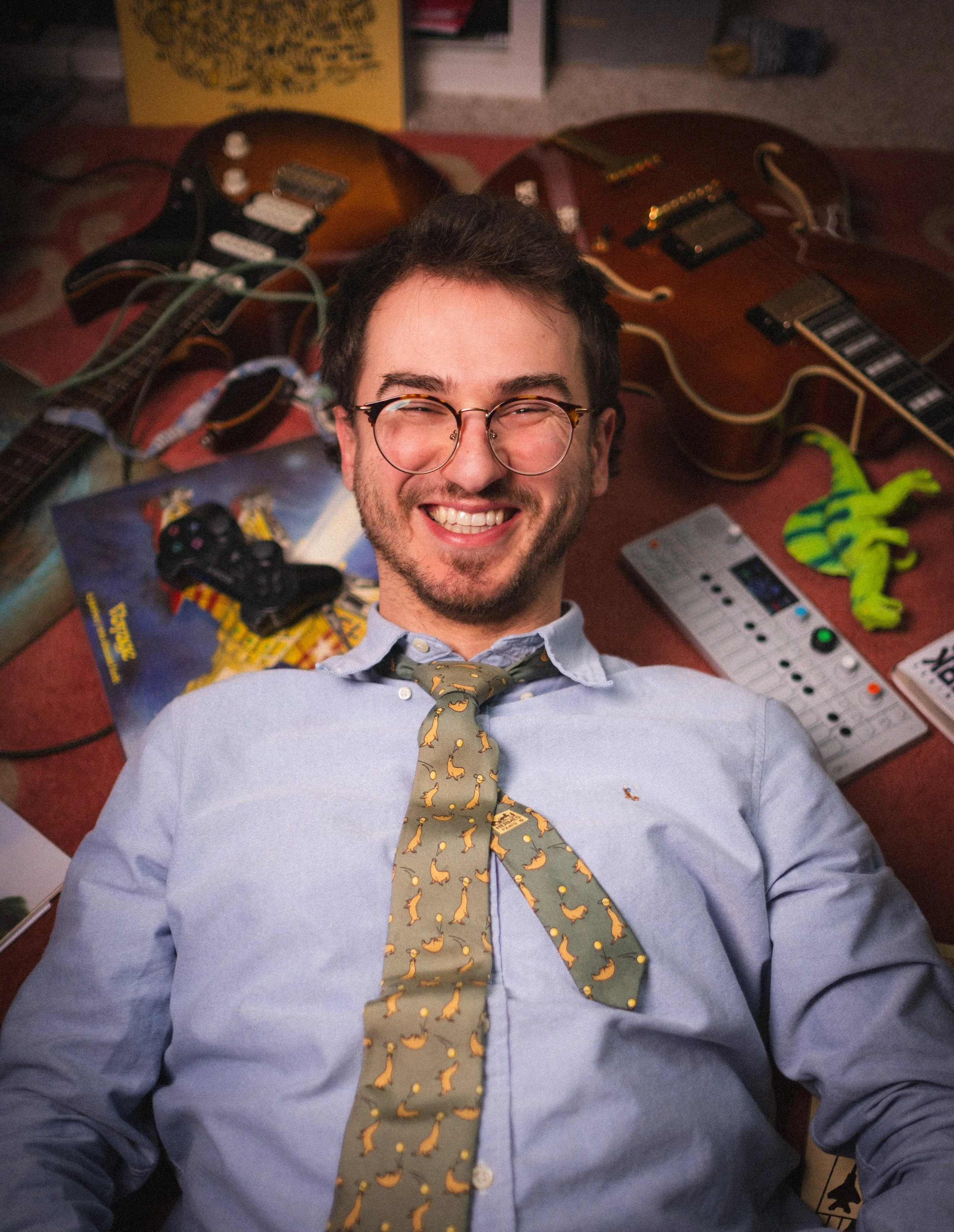 Un homme souriant, avec des lunettes, portant une chemise bleue et une cravate à motifs de petits canards, allongé sur un lit avec des guitares, un synthétiseur, une console de jeux et un jouet en forme de lézard en arrière-plan.