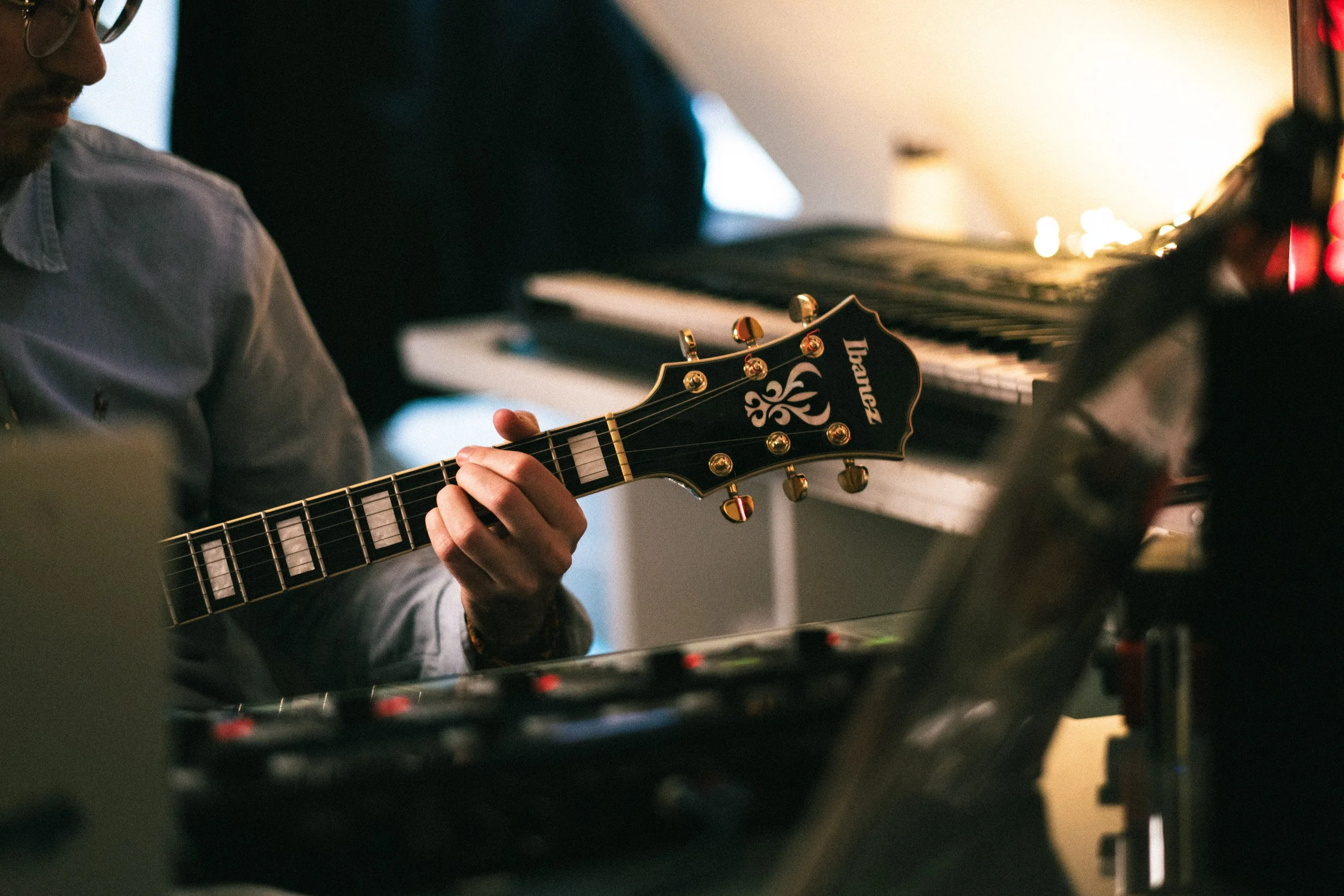 Une personne joue de la guitare électrique dans une pièce avec un piano en arrière-plan, lumières tamisées.