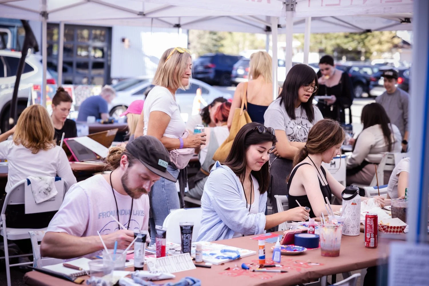 People participating in an outdoor art and crafts event under a white canopy, with tables covered in various art supplies and drinks, in a parking lot with cars in the background.