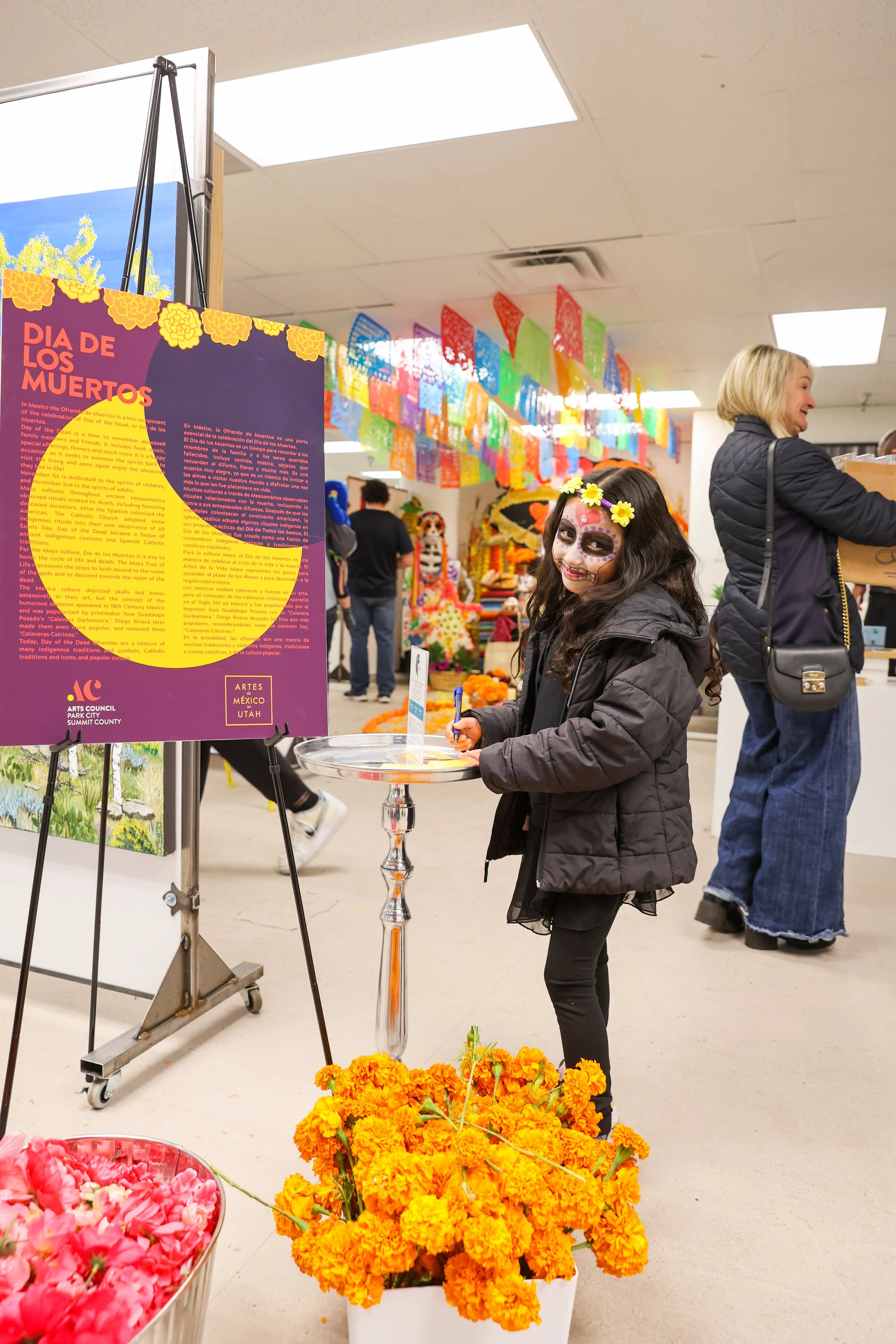 Menina com o rosto pintado para o Dia de los Muertos, usando flores no cabelo, ao lado de um cartaz informativo, numa celebração interna decorada com bandeiras coloridas de papel picado e flores de calêndula.