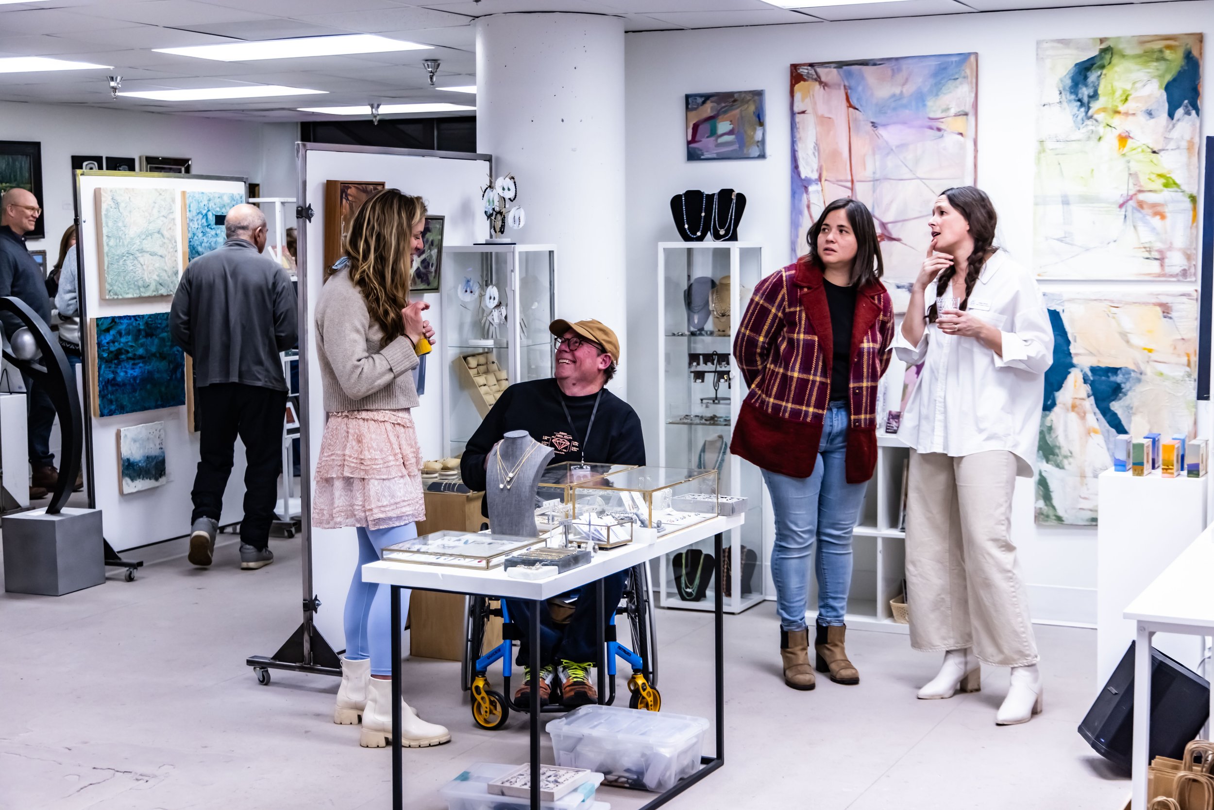 People shopping at an art and jewelry gallery, some looking at paintings and jewelry displays, engaged in conversations.