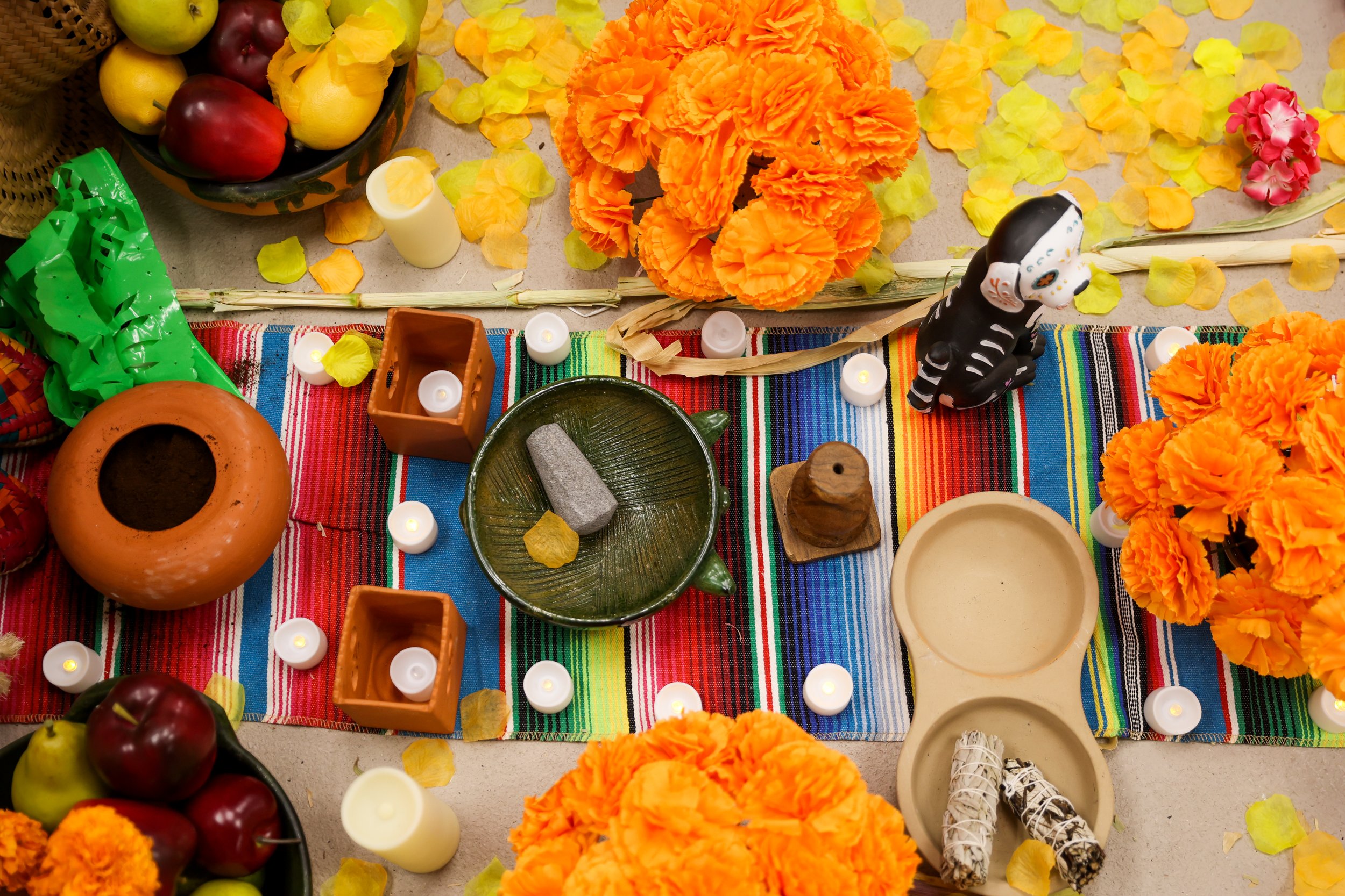 Um altar colorido tradicional mexicano do Dia dos Mortos com flores de calêndula, velas, maçãs, uma cesta verde, uma estatueta de caveira e itens decorativos sobre um pano listrado.