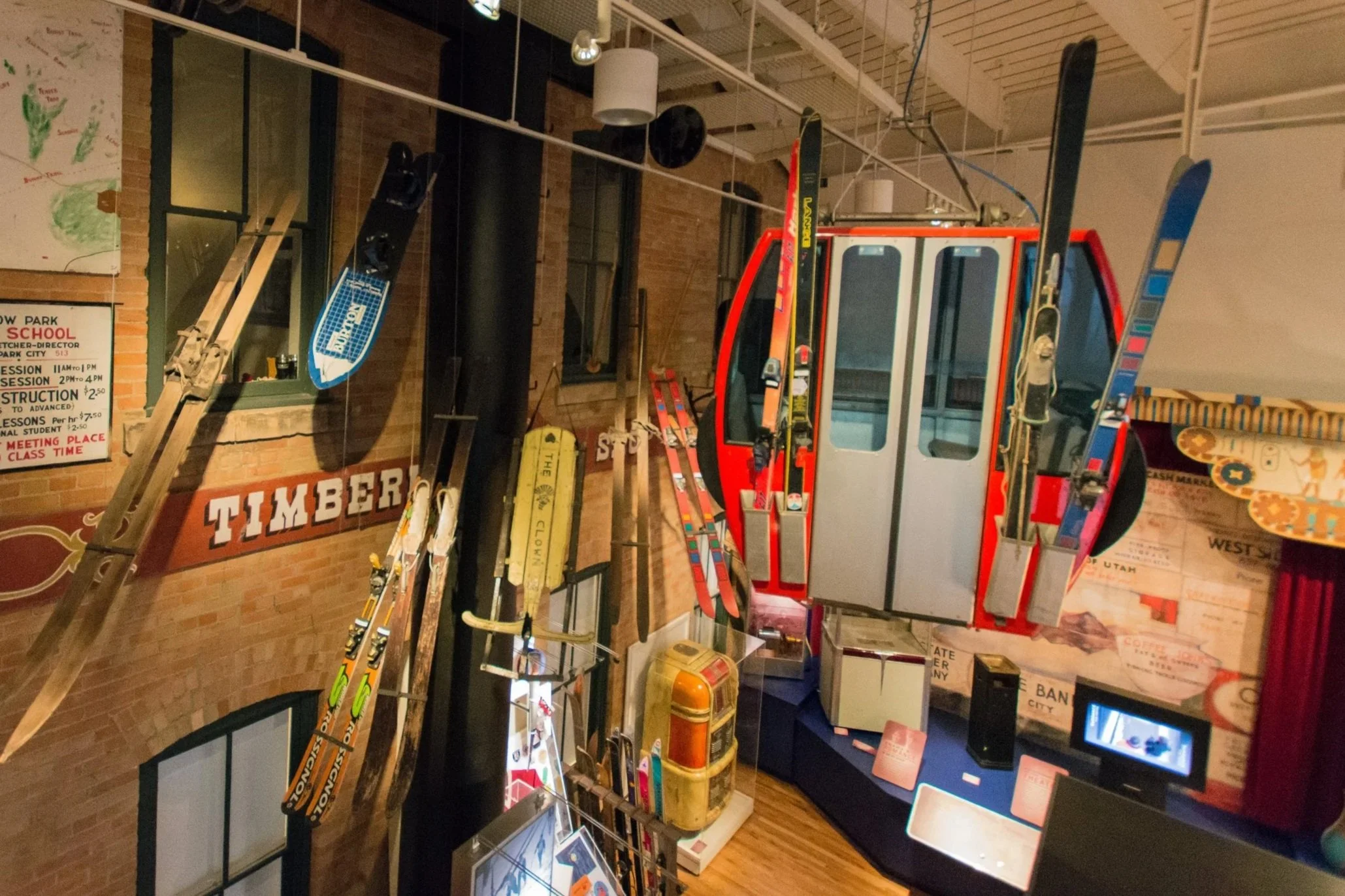 Indoor museum display with vintage skis hanging from the ceiling, a red gondola, ski equipment, and a mural with maps and a sign that says 'Timber'.