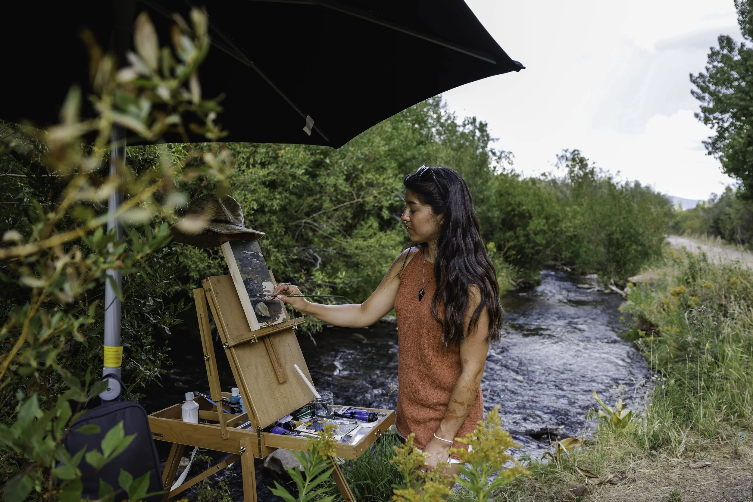 Mulher a pintar ao ar livre num pequeno cavalete junto a um rio, com um guarda-chuva preto para se proteger do sol, rodeada por vegetação.