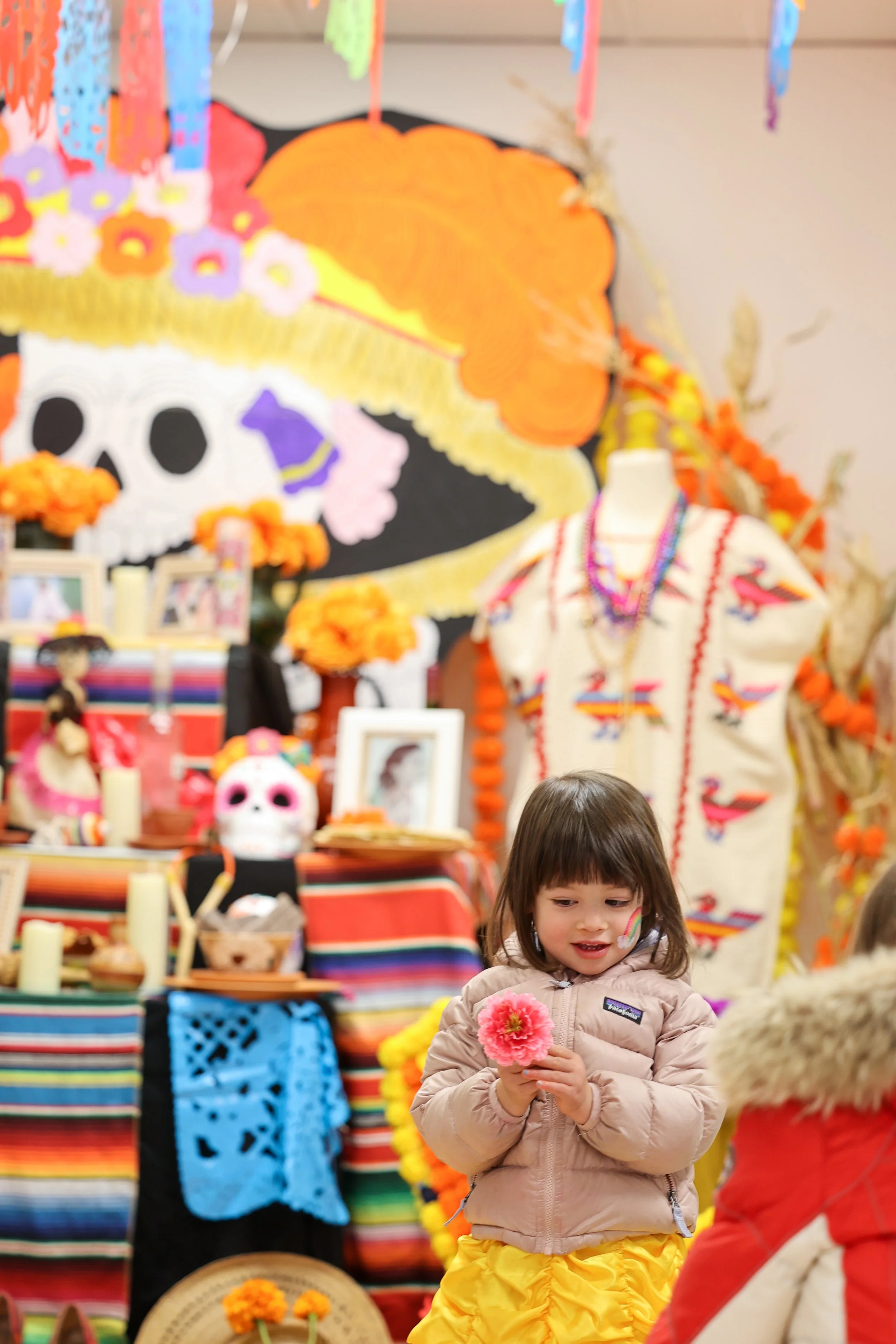 A young girl holding a pink flower at a Day of the Dead celebration with vibrant Mexican decorations, including a large skull mural with colorful flowers, marigold garlands, and traditional Mexican textiles.