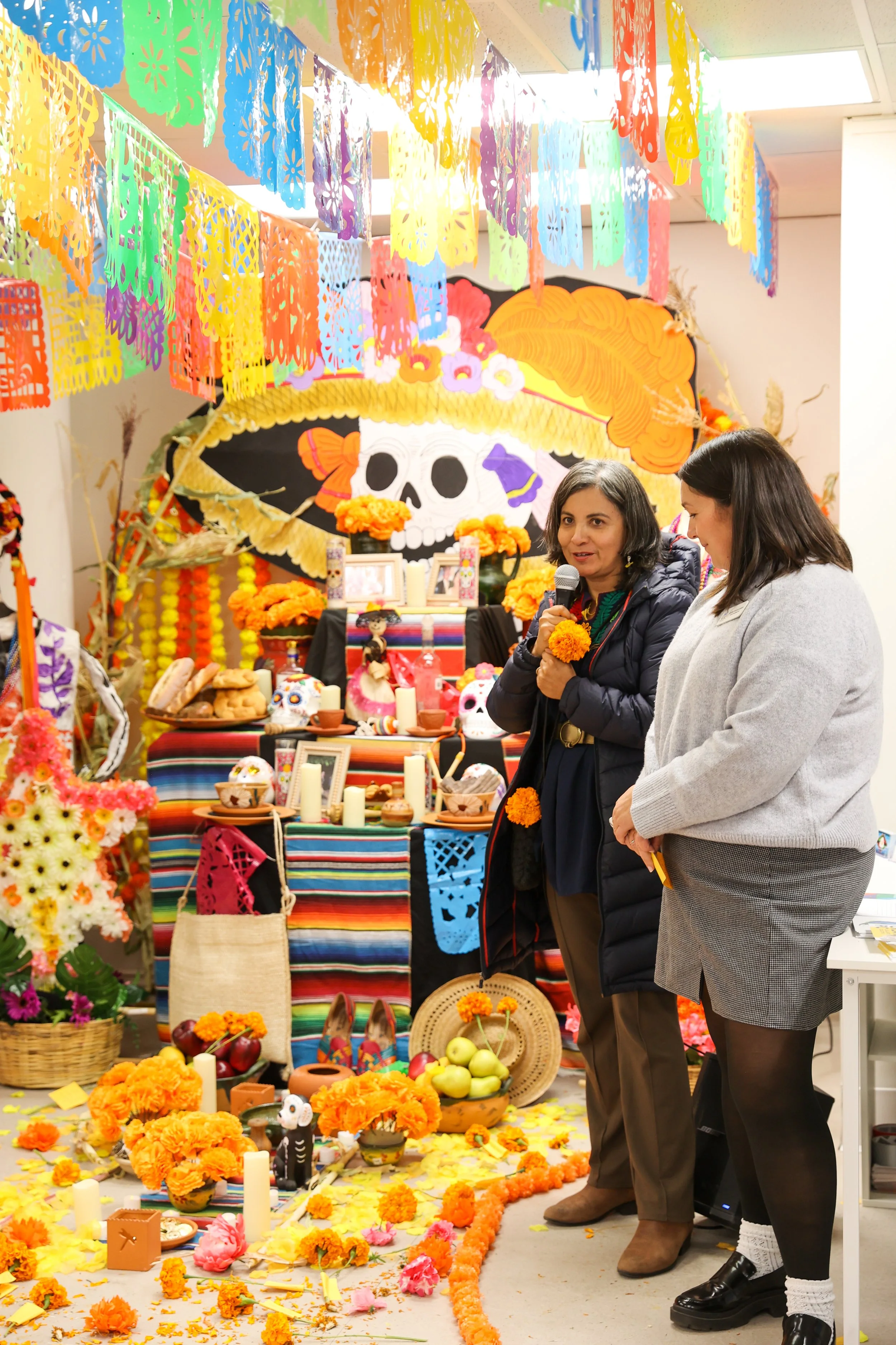 Celebração do Dia de los Muertos com bandeiras coloridas de papel picado, altar tradicional decorado com malmequeres, velas, fotos e caveiras de açúcar, com mulheres a conversar em meio a decorações vibrantes.