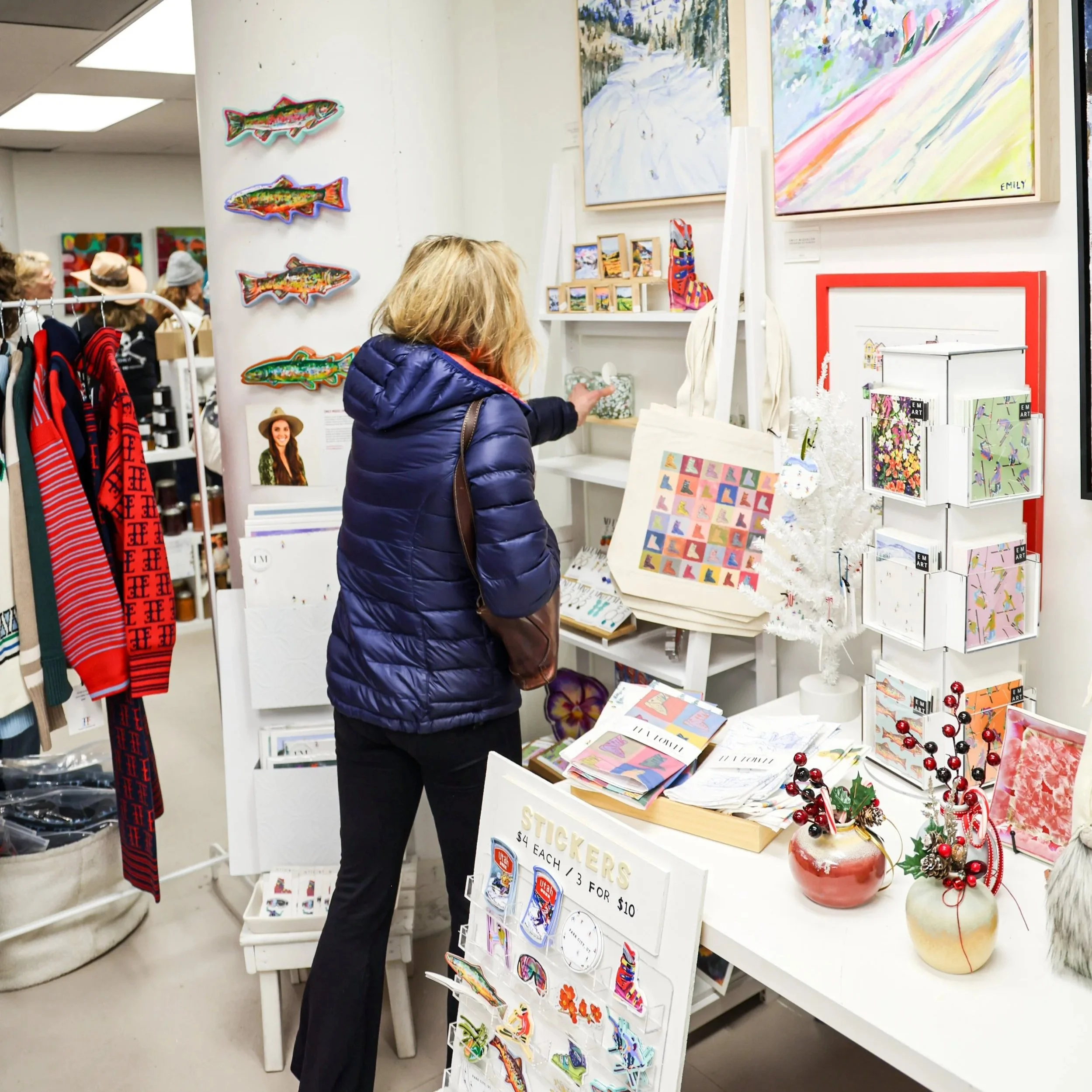 A woman shopping at an art and gift store, wearing a blue jacket, with various colorful art, holiday decorations, and stationery on display around her.