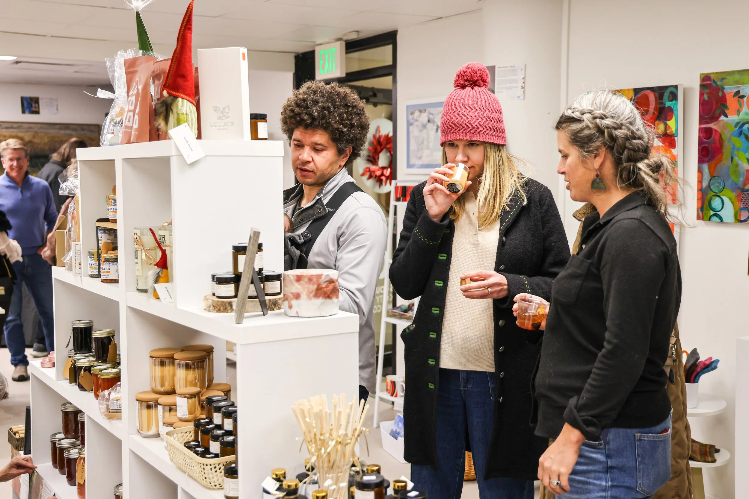 People shopping at a store, looking at products on white shelves. Three women in the foreground are tasting or sniffing items, one wearing a red knit hat, and a man behind them appears to be assisting.