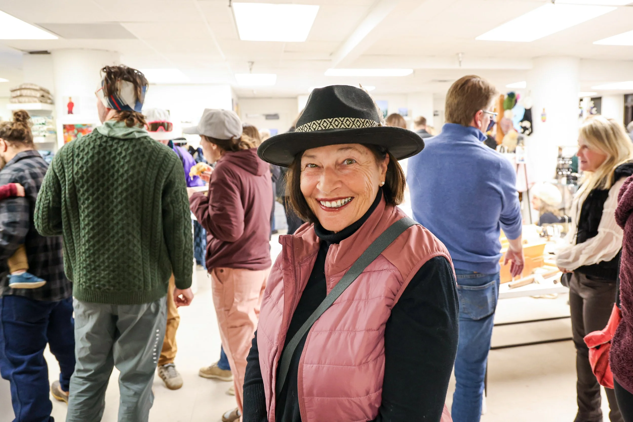 A woman smiling and wearing a black wide-brimmed hat with a decorative band, pink vest, and black shirt, in a crowded indoor shopping or market area.