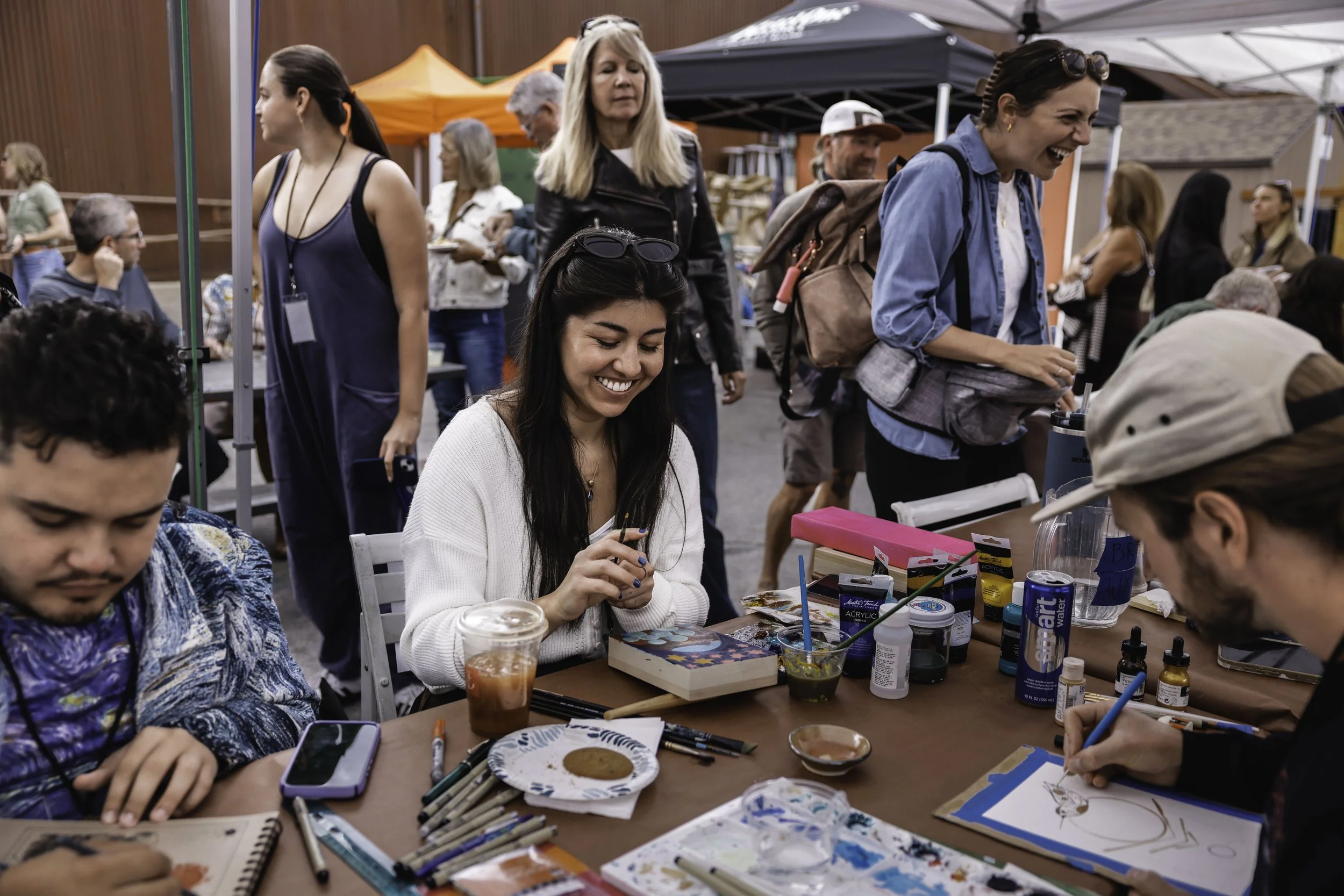 People attending an art event outdoors, with some participants drawing or painting at tables and others socializing in the background.