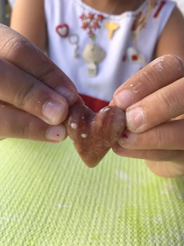 Child holding a heart-shaped stone or object with a reddish-brown color and white spots, with a green textured surface underneath and a blurred background of a person wearing a white shirt with colorful embroidered patches.
