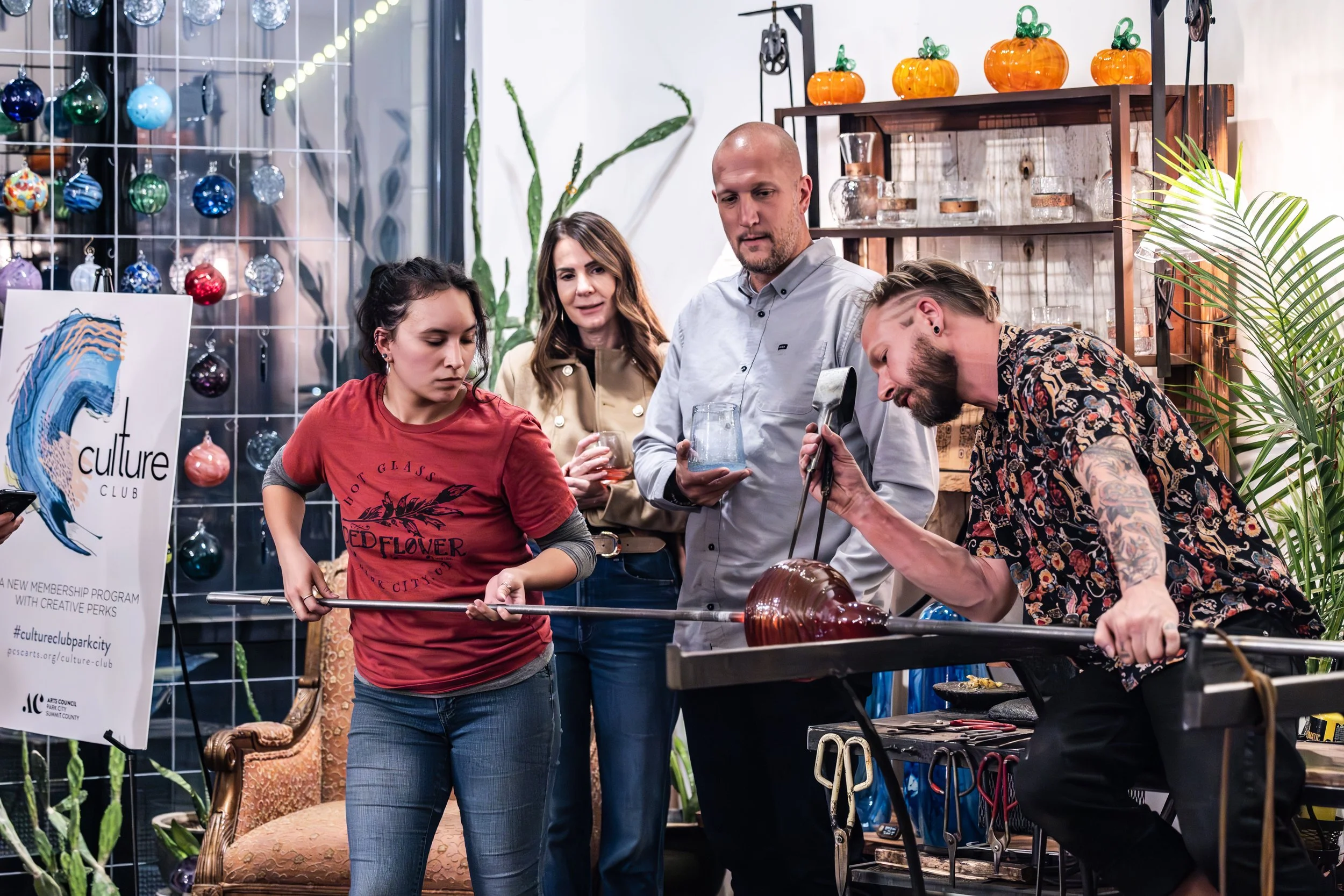 Glass artist shaping molten glass using a blowpipe at a workshop, with three women observing, in a decorated studio with plants, shelves, and ornaments.