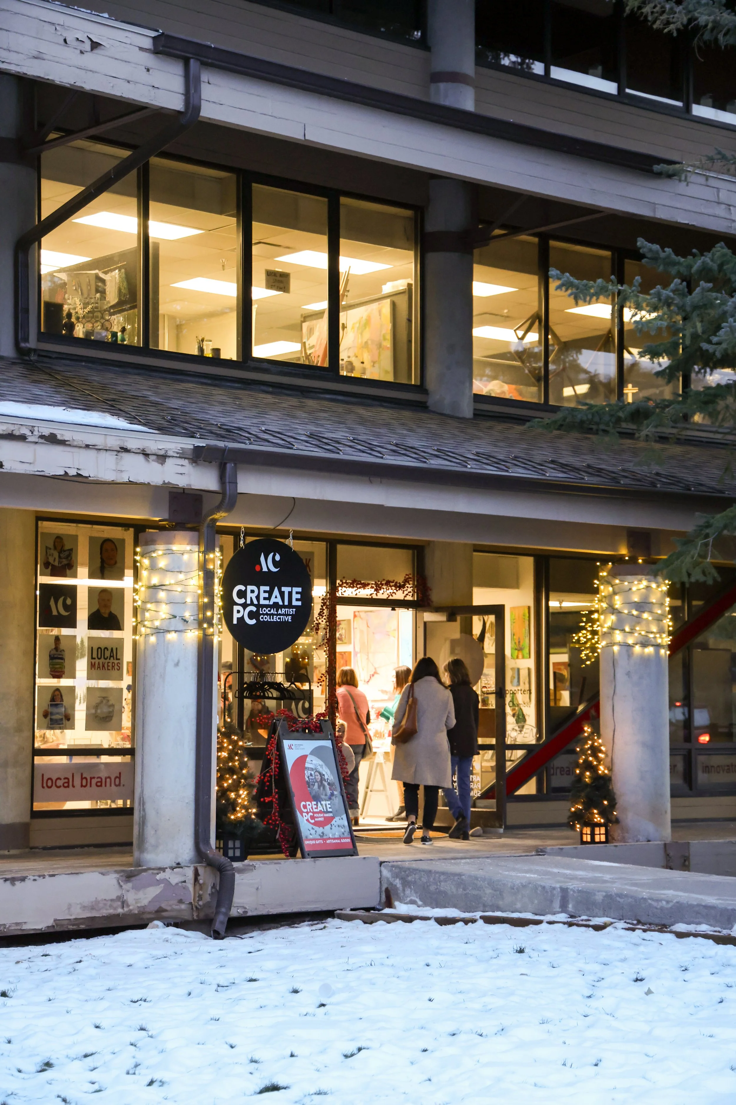 People entering a shop decorated with string lights and Christmas trees, with snow on the ground outside.