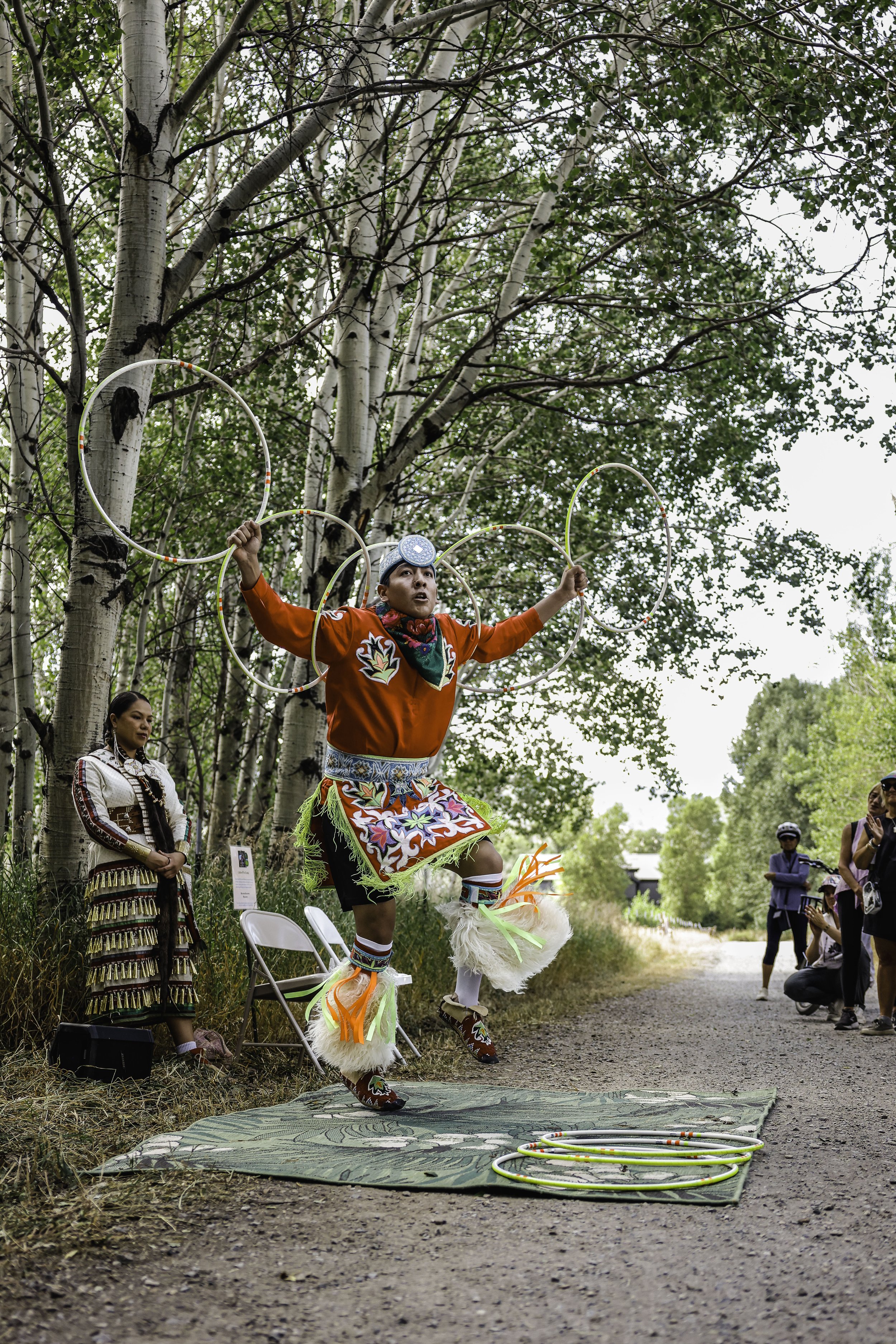 A Native American man in traditional attire performing a hoop dance outdoors on a dirt path, with spectators watching. The dancer is balancing multiple hoops and wearing colorful clothing and accessories, including fur shoes and beadwork, surrounded by trees.