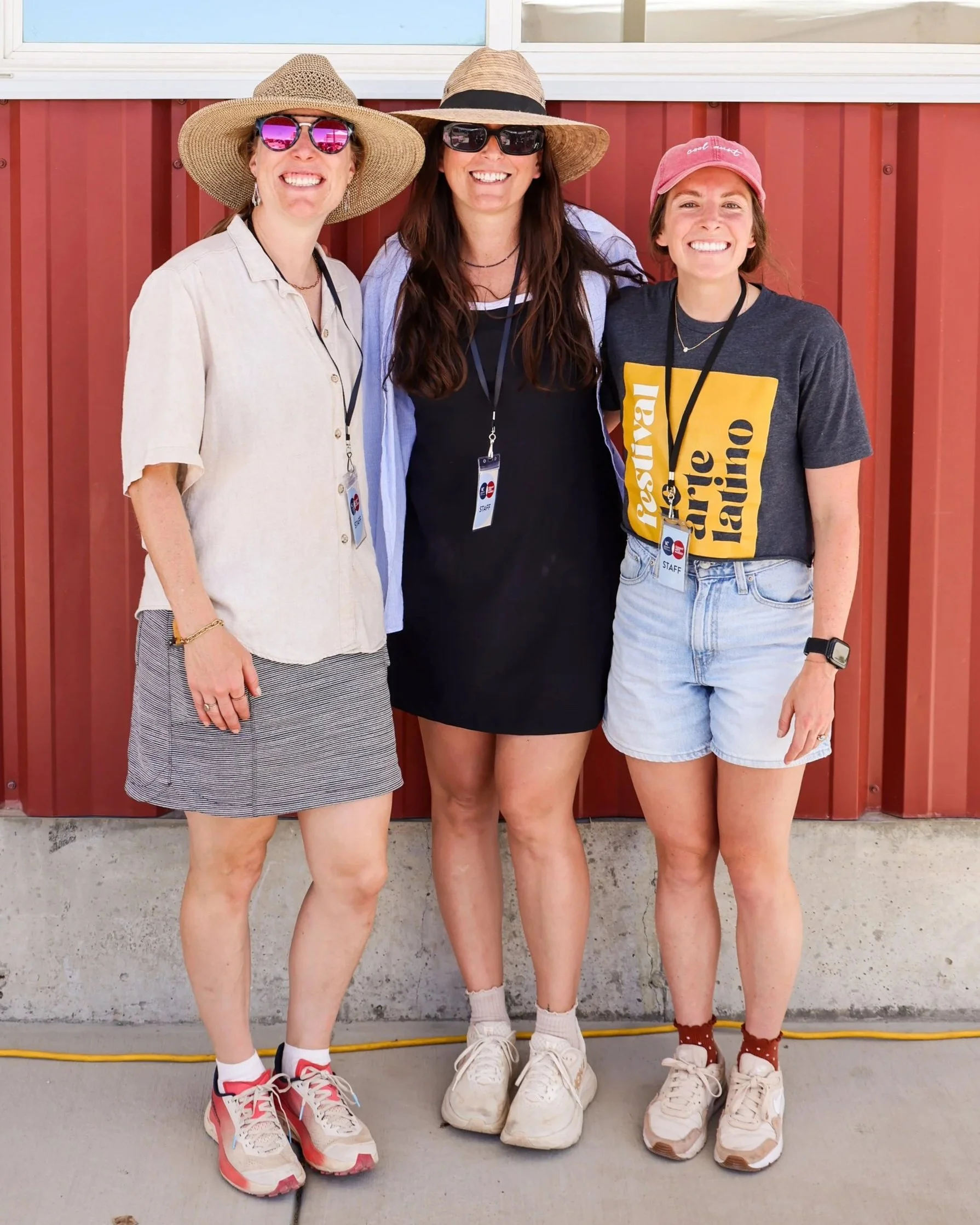 Three women standing side by side in front of a red wall, smiling at the camera. They are wearing sunglasses, hats, casual clothing, and sneakers, with lanyards hanging around their necks.