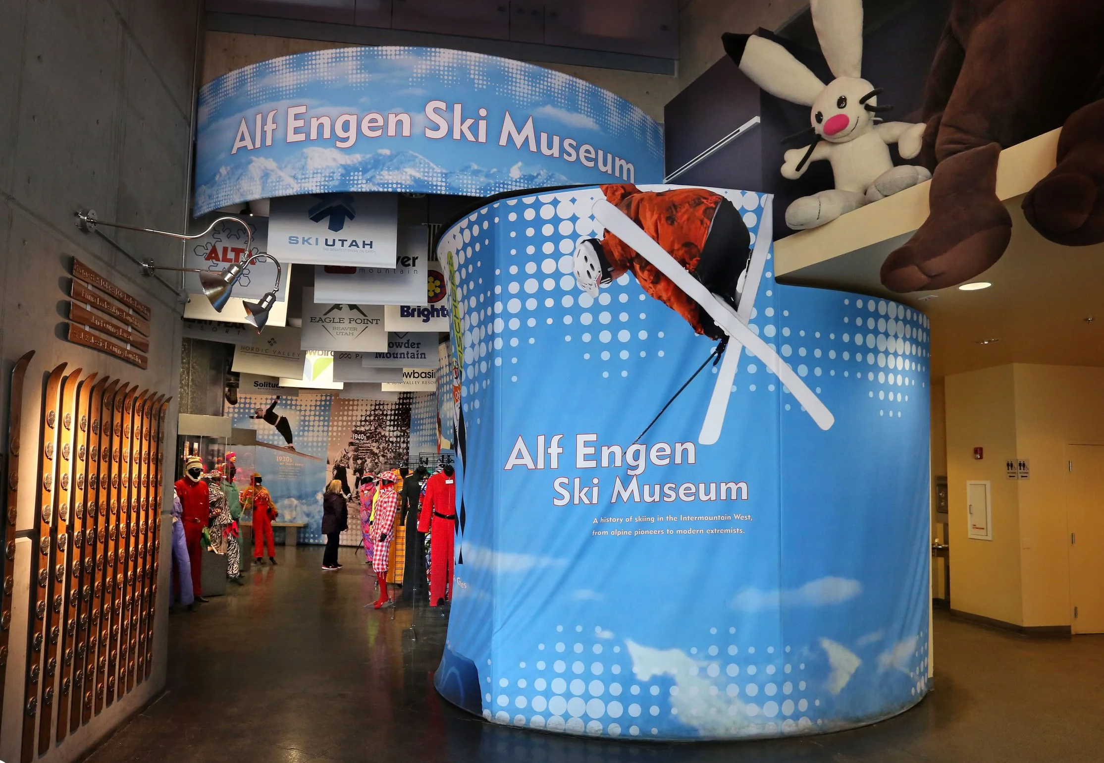 Entrance to the Alf Engen Ski Museum with a curved blue banner featuring a skier graphic and text, plush rabbit toy and brown boots hanging overhead, mannequins in colorful ski suits inside, wooden plaques on the wall, and people exploring the museum.