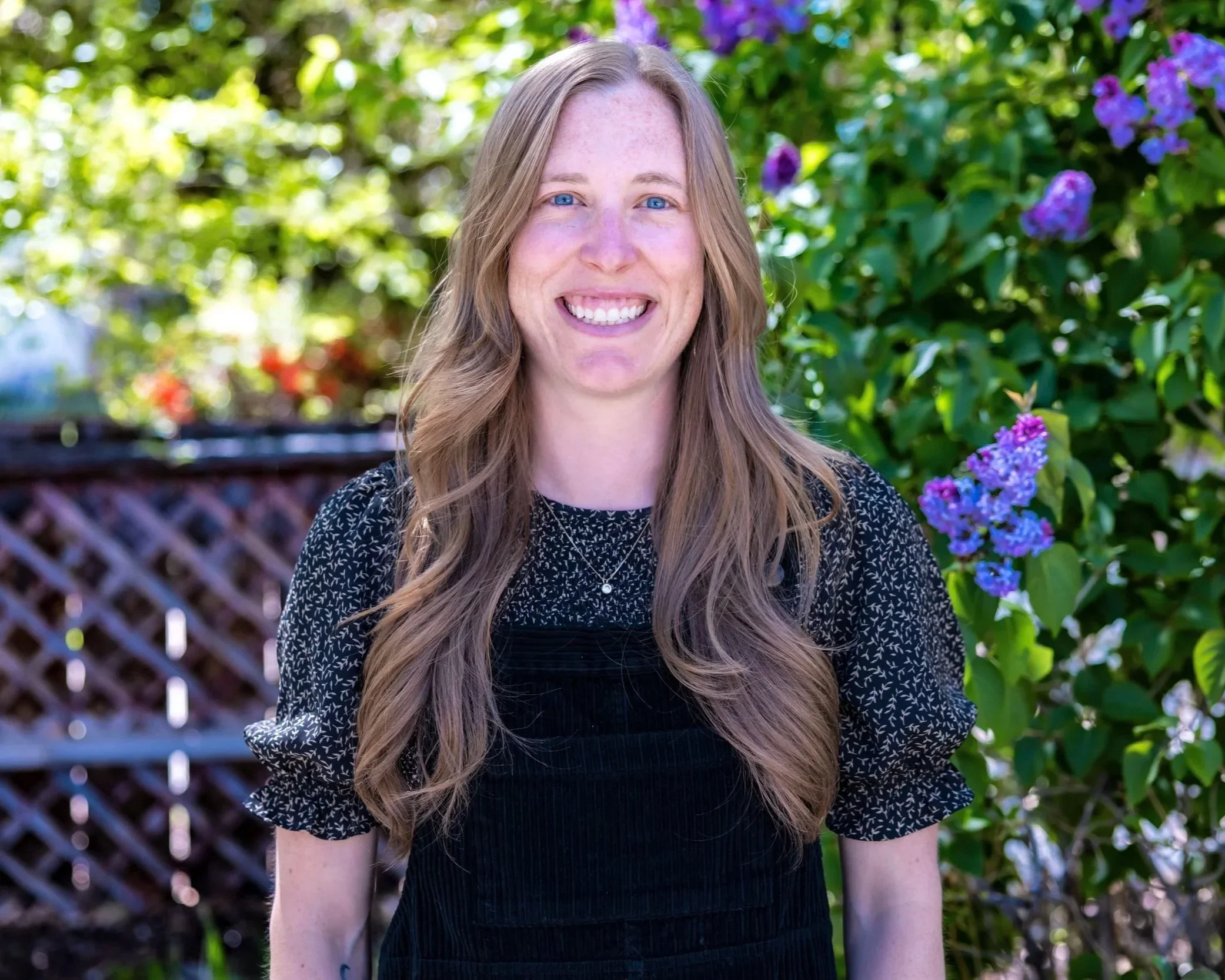 A smiling woman with long, wavy light brown hair standing outdoors in front of green foliage and purple flowers.