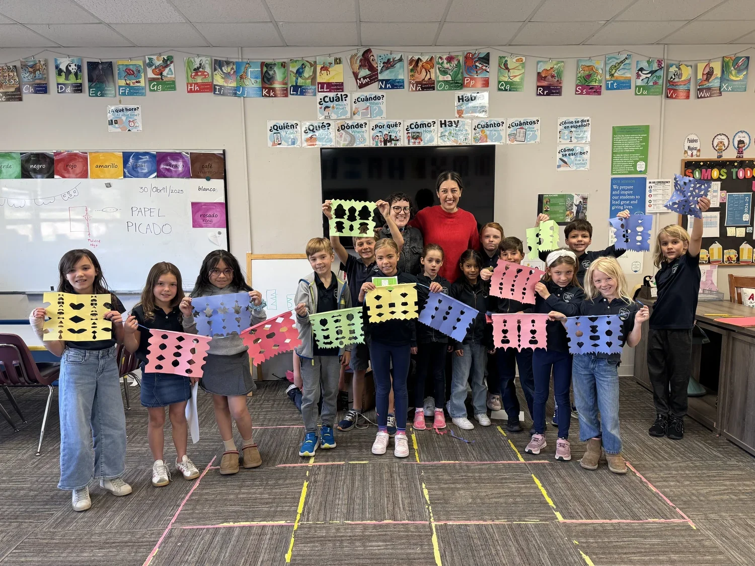 A group of children in a classroom holding colorful paper banners with cut-out shapes, smiling for the photo with two teachers behind them. The classroom has educational decorations and a whiteboard with writing.