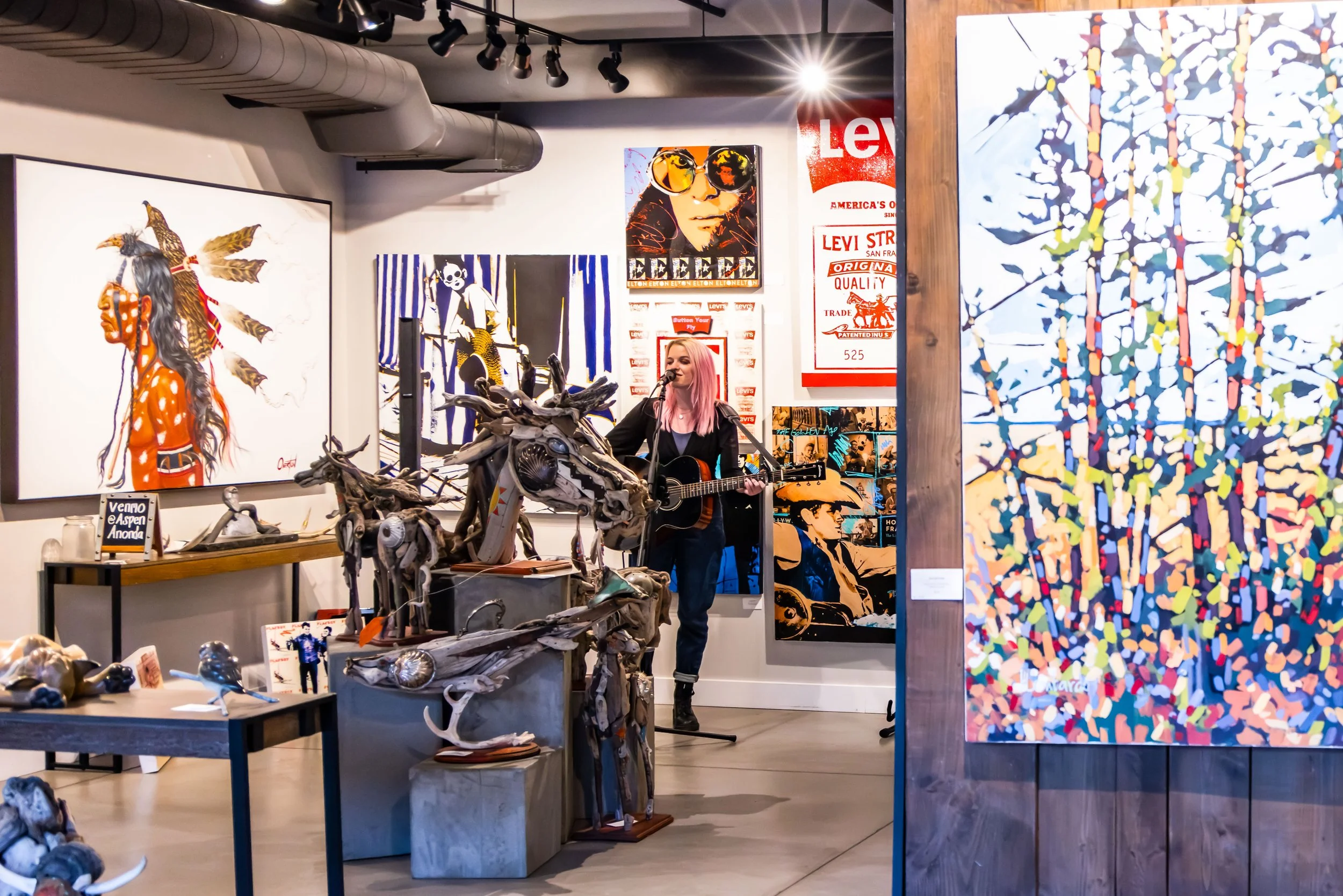 A woman with pink hair playing guitar and singing in an art gallery with various paintings, sculptures, and posters on the walls and display tables.