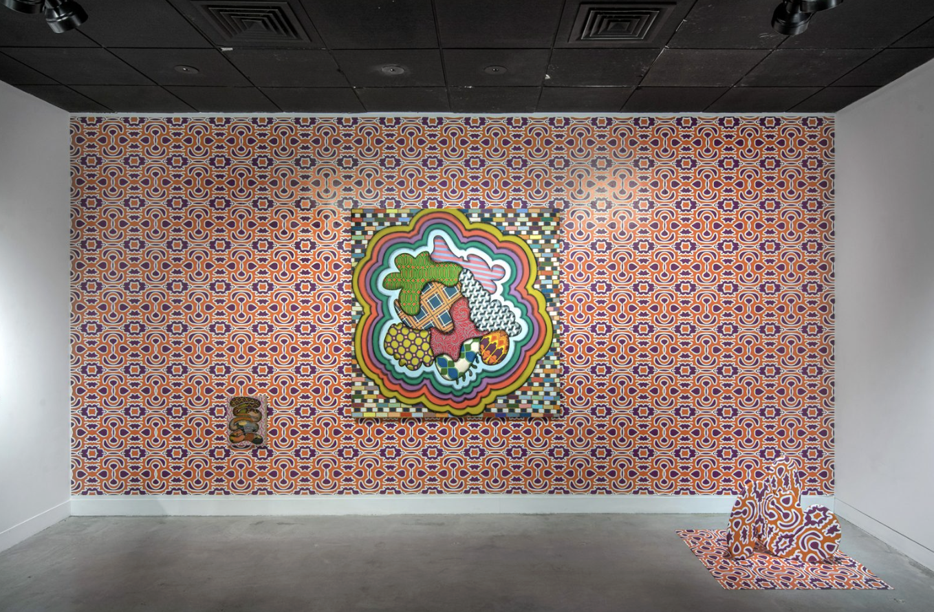 Uma instalação artística apresenta um papel de parede vibrante e estampado cobrindo toda uma parede, com uma obra de arte colorida e multicolorida montada sobre ele. A obra de arte inclui várias formas e padrões, rodeados por um fundo xadrez. À direita, há uma peça com padrão correspondente no chão e, à esquerda, uma pequena escultura ou objeto também está colocado na parede.