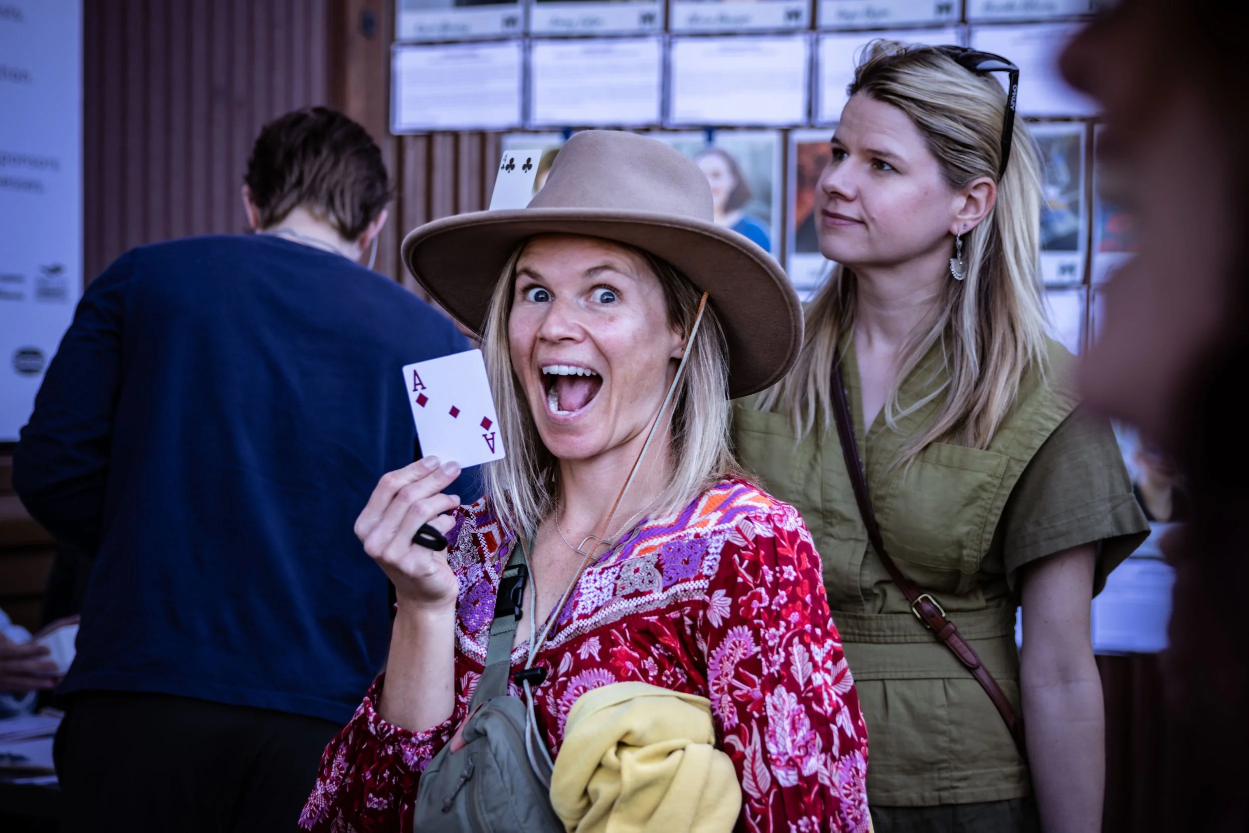 A woman wearing a hat and a colorful patterned top holding an ace of diamonds playing card, smiling widely at the camera in a crowded indoor setting.