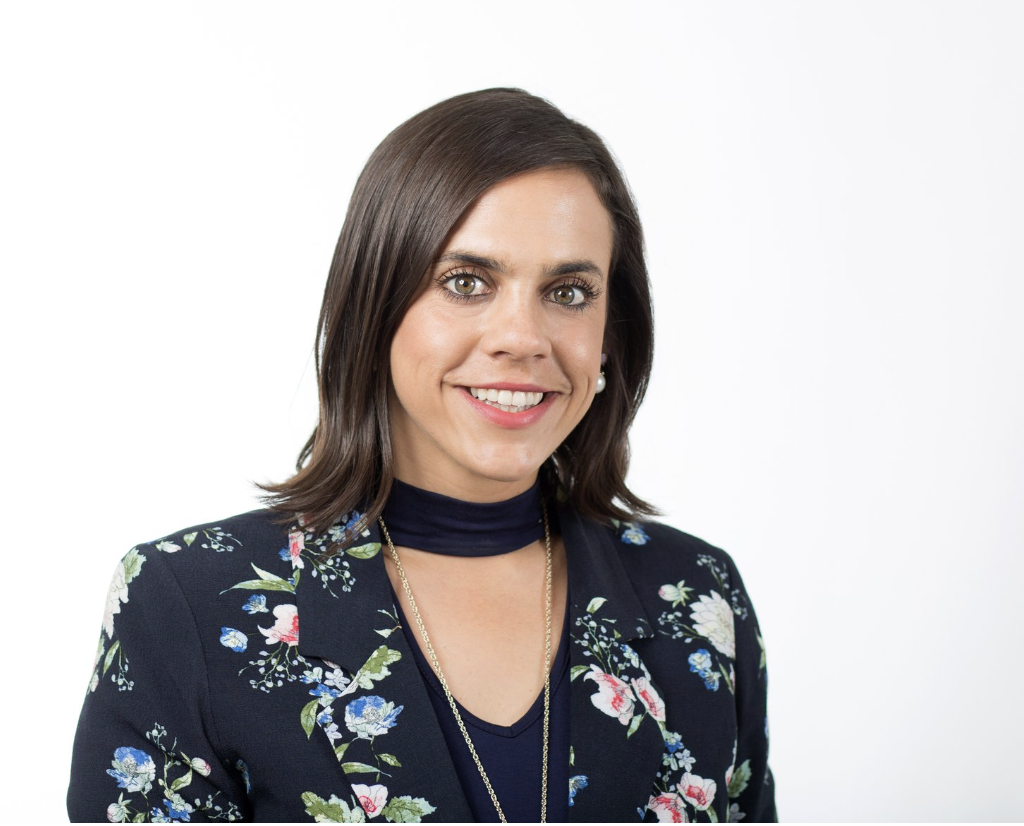 A woman with shoulder-length brown hair, wearing a navy floral blazer and a navy turtleneck, smiling against a white background.