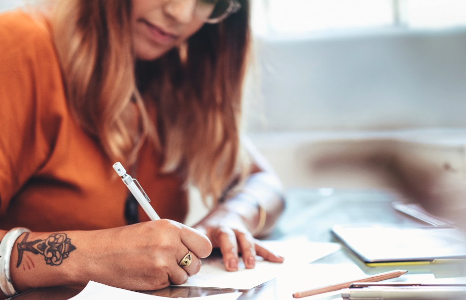 Uma mulher com tatuagens no braço a escrever em papel com uma caneta numa secretária.