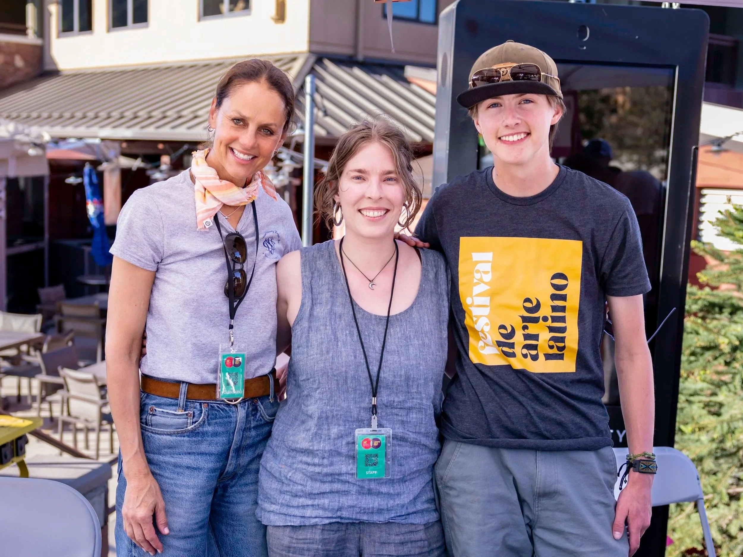 Three smiling people standing outdoors in a social gathering, with a woman on the left, a woman in the middle, and a young man on the right. All are wearing casual clothing and have event badges around their necks.